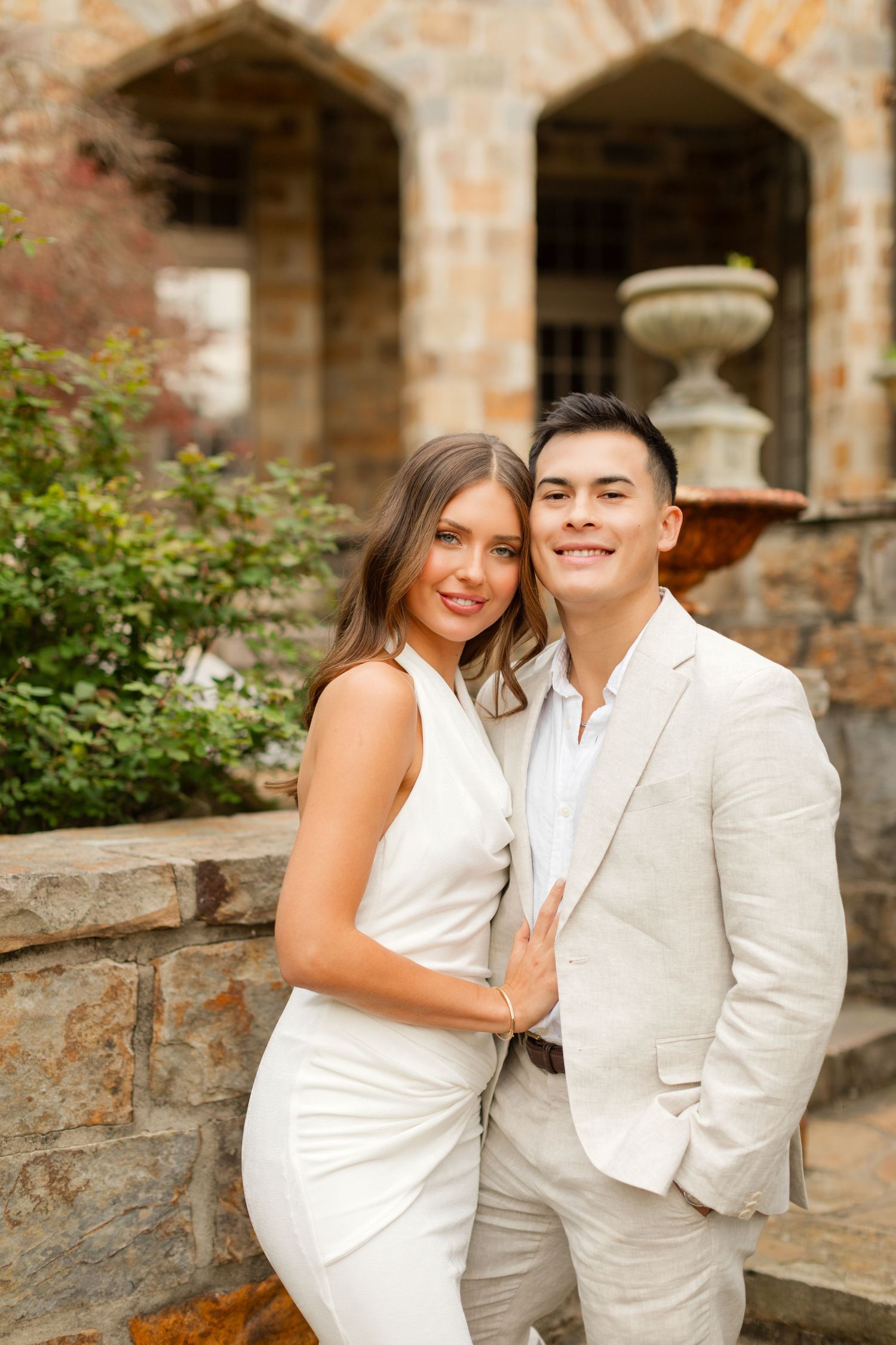 A couple in white and beige attire poses together against a rustic stone wall with greenery. Goodwin Manor.