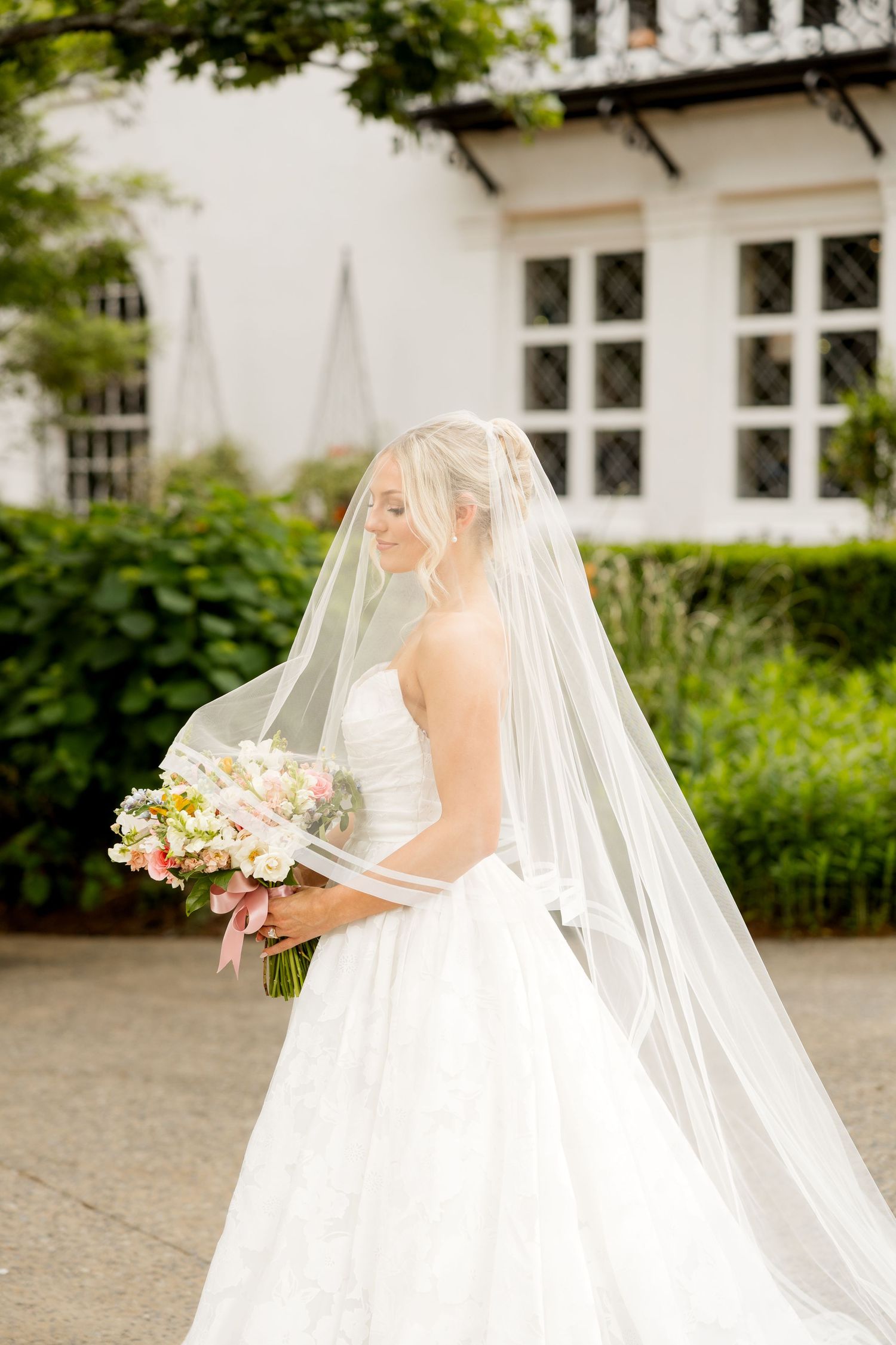 A bride in a flowing white wedding gown and long veil stands in a garden courtyard holding a bouquet.