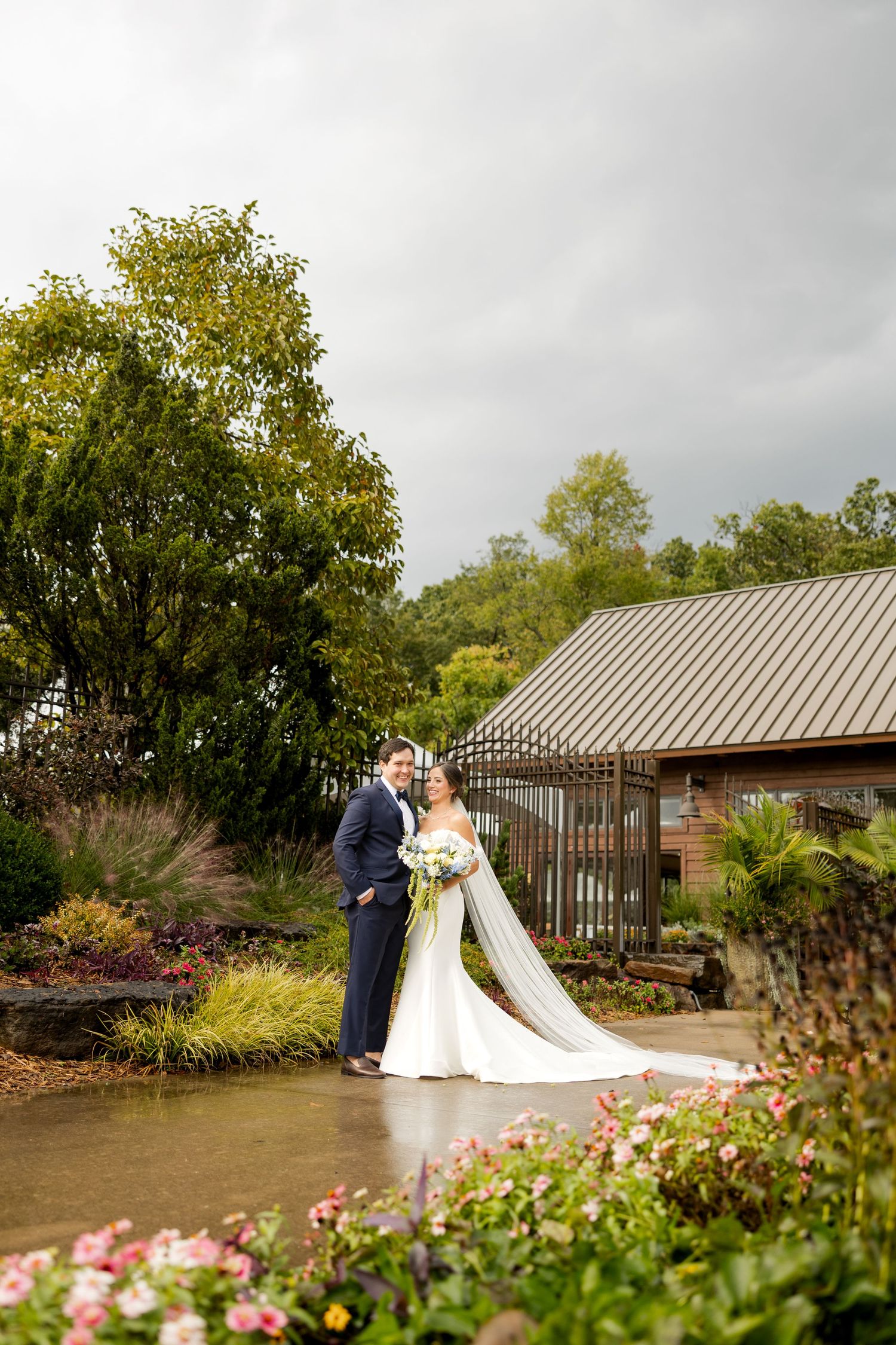 A wedding couple poses in front of a rustic barn with lush gardens and flowering plants on an overcast day.Botanical Gardens of the Ozarks