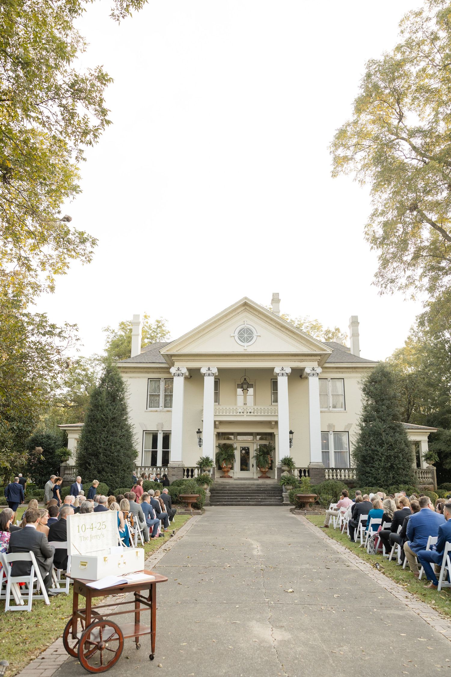 An elegant white mansion with tall columns hosts an outdoor wedding ceremony with guests seated along a stone pathway.