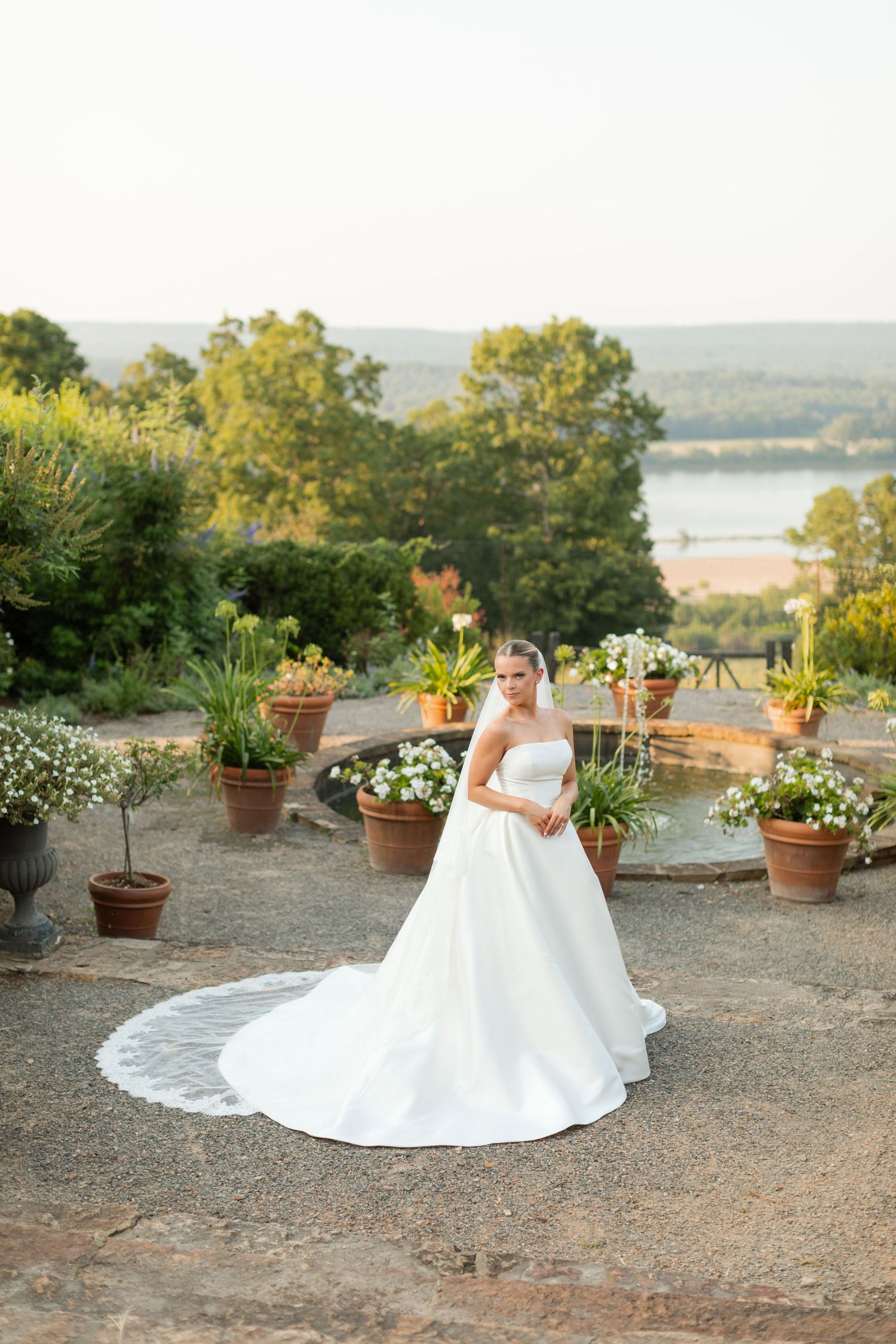 A white wedding dress flows elegantly against a scenic overlook with potted flowers and greenery at sunset.