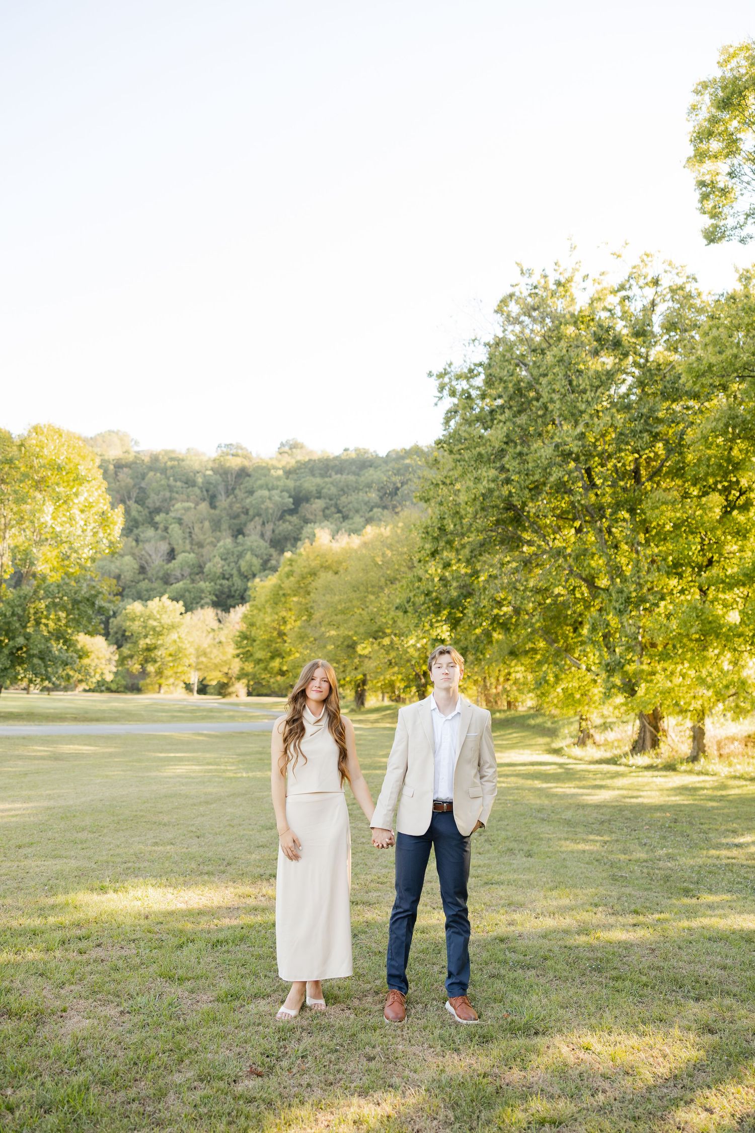 Couple walking together in a sunny meadow during golden hour with trees in the background.