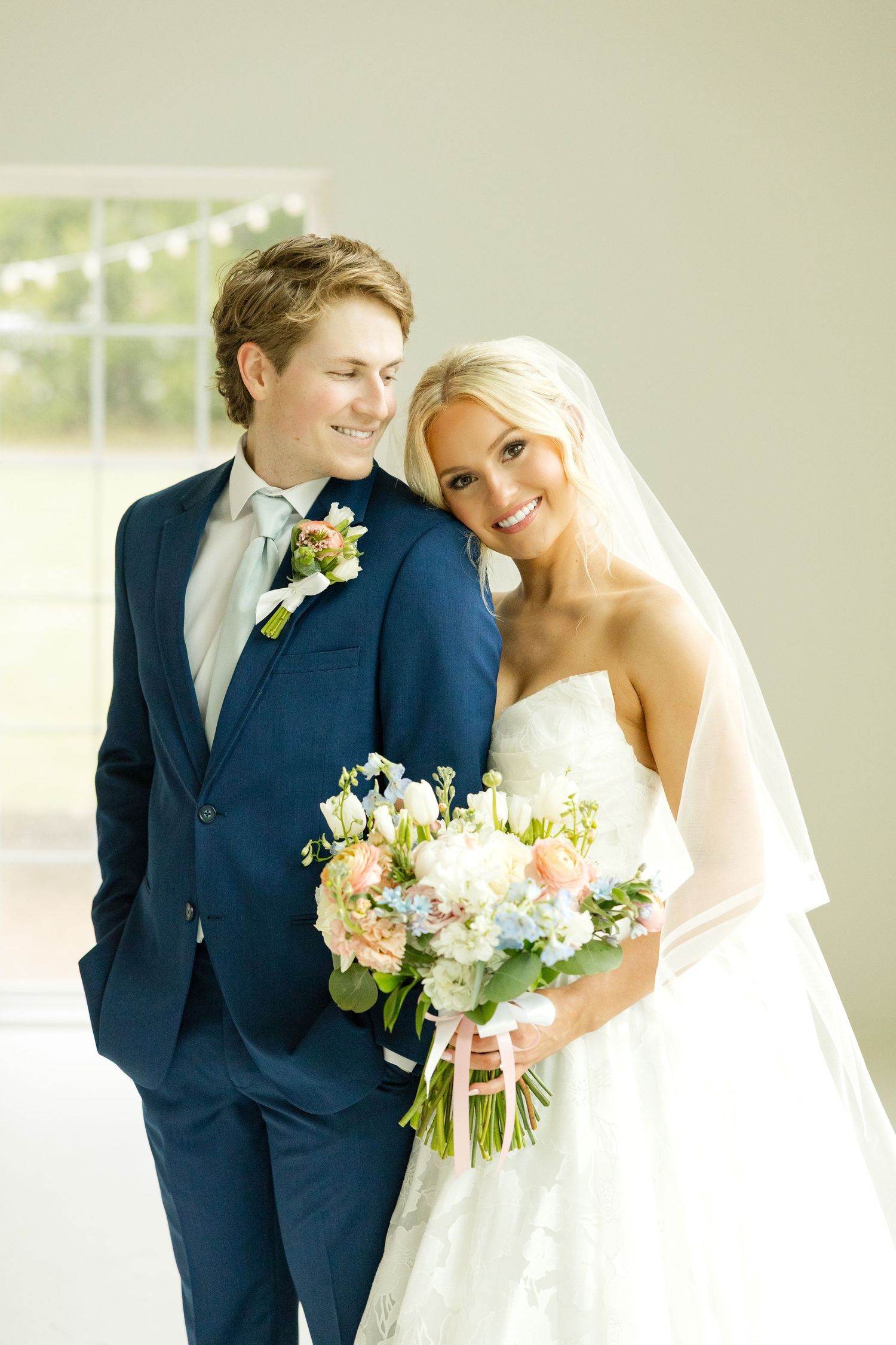 A couple on their wedding day wearing a navy suit and white gown, holding a pastel floral bouquet.