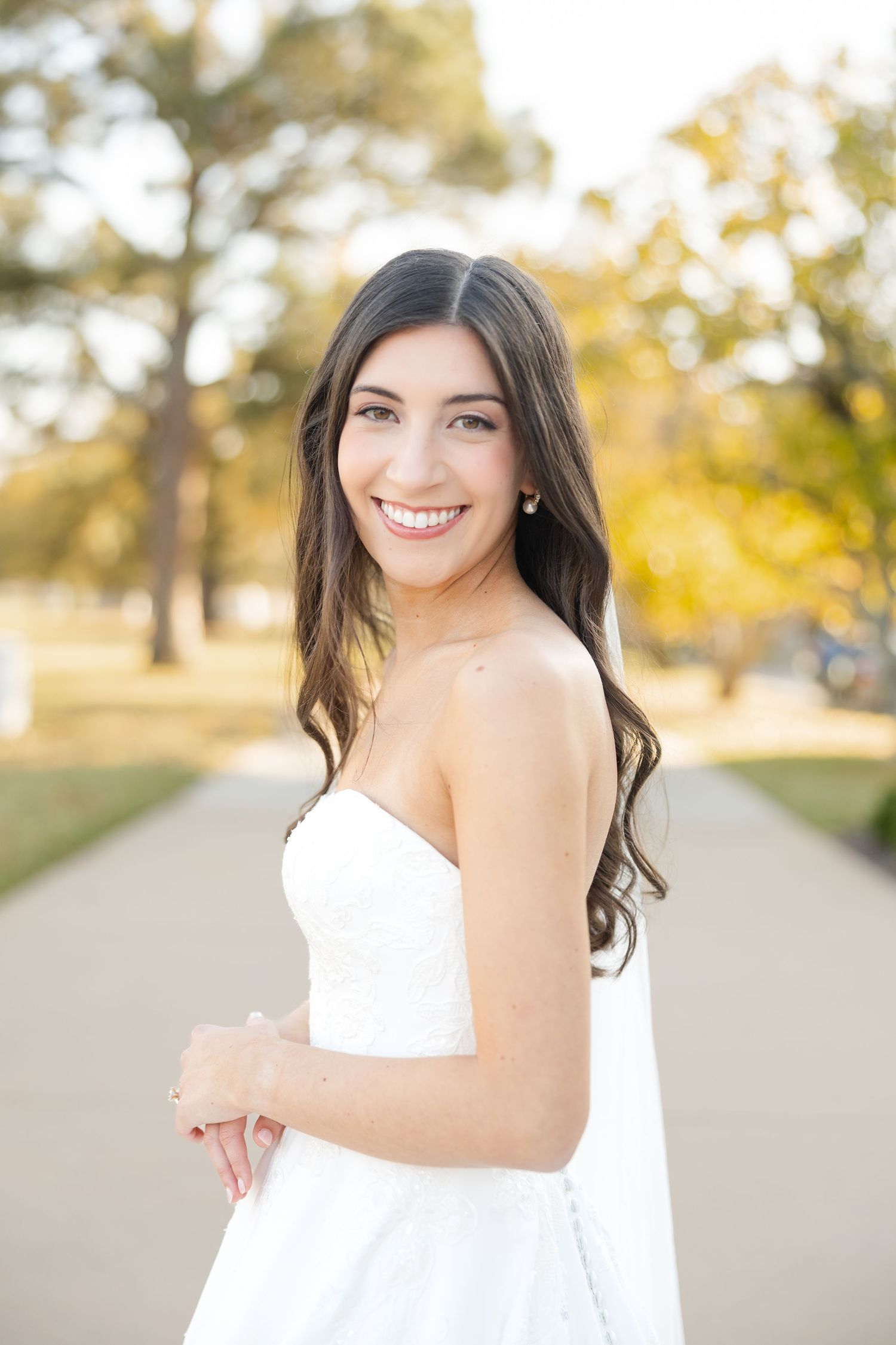 A strapless white wedding gown shimmers in warm sunlight against a backdrop of fall trees.