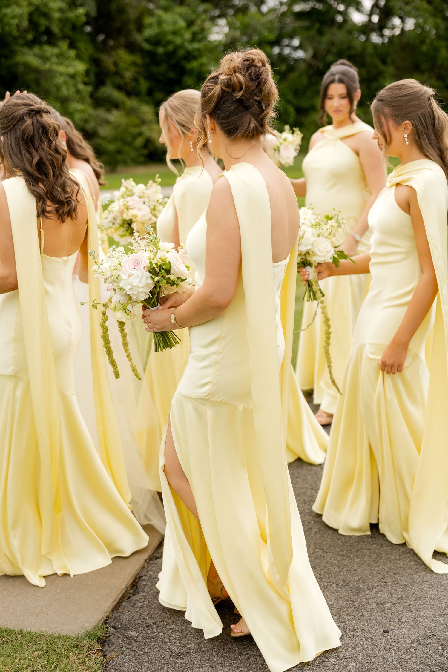 Bridesmaids in matching pale yellow gowns holding white floral bouquets walk together outdoors at a wedding.