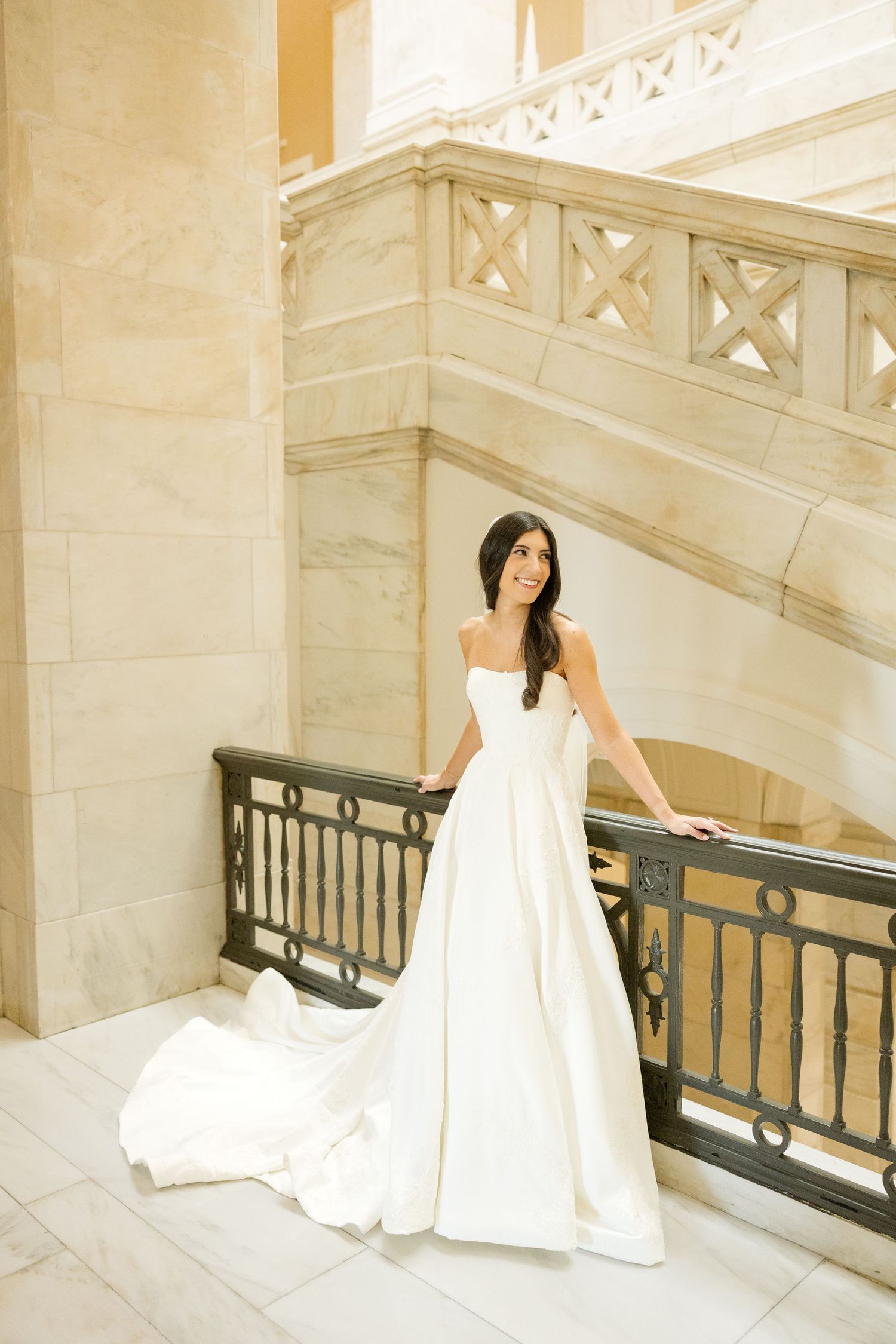 A bride in a white wedding gown poses elegantly on a grand marble staircase with ornate railings.