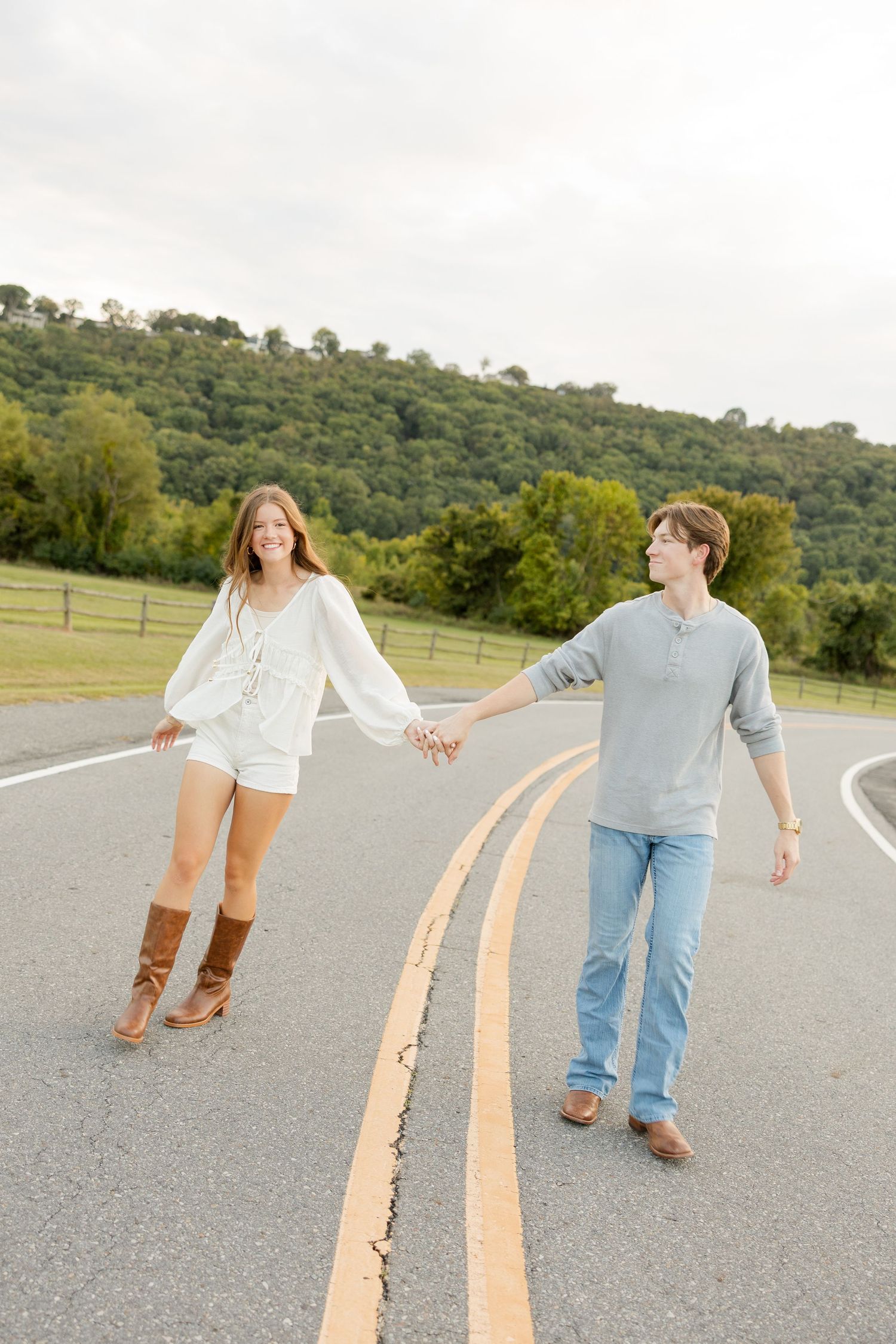 A couple walks along yellow road lines holding hands with a hillside in the background.