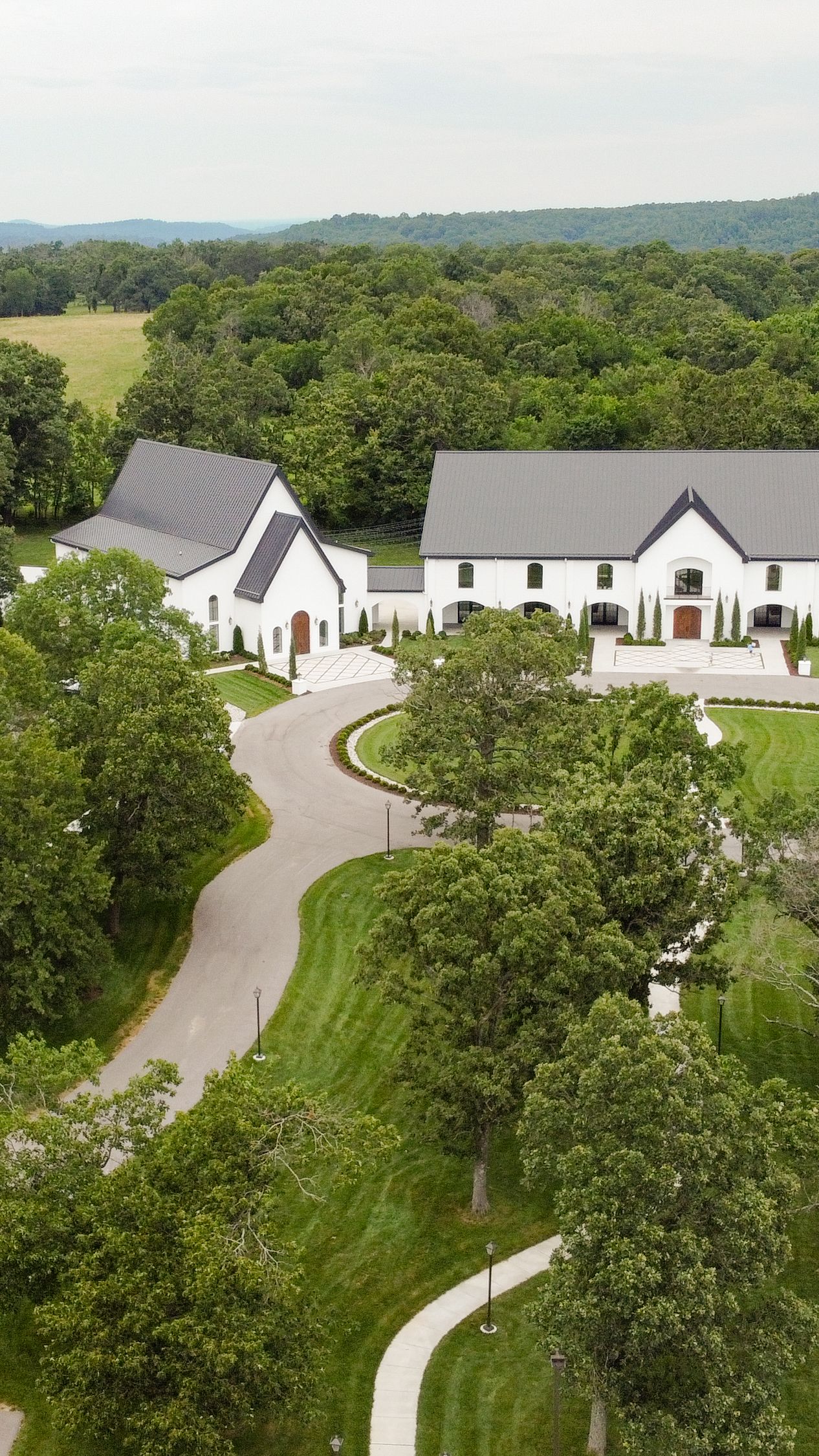 Aerial view of modern white residential buildings with gray roofs arranged along a curved driveway surrounded by lush trees.