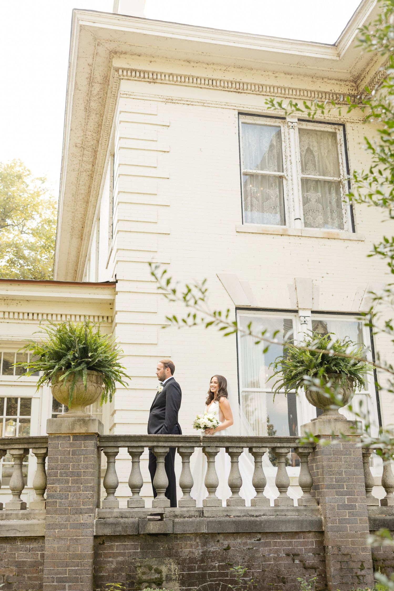 Elegant wedding couple walks along ornate stone balustrade outside historic mansion with white columns and greenery.