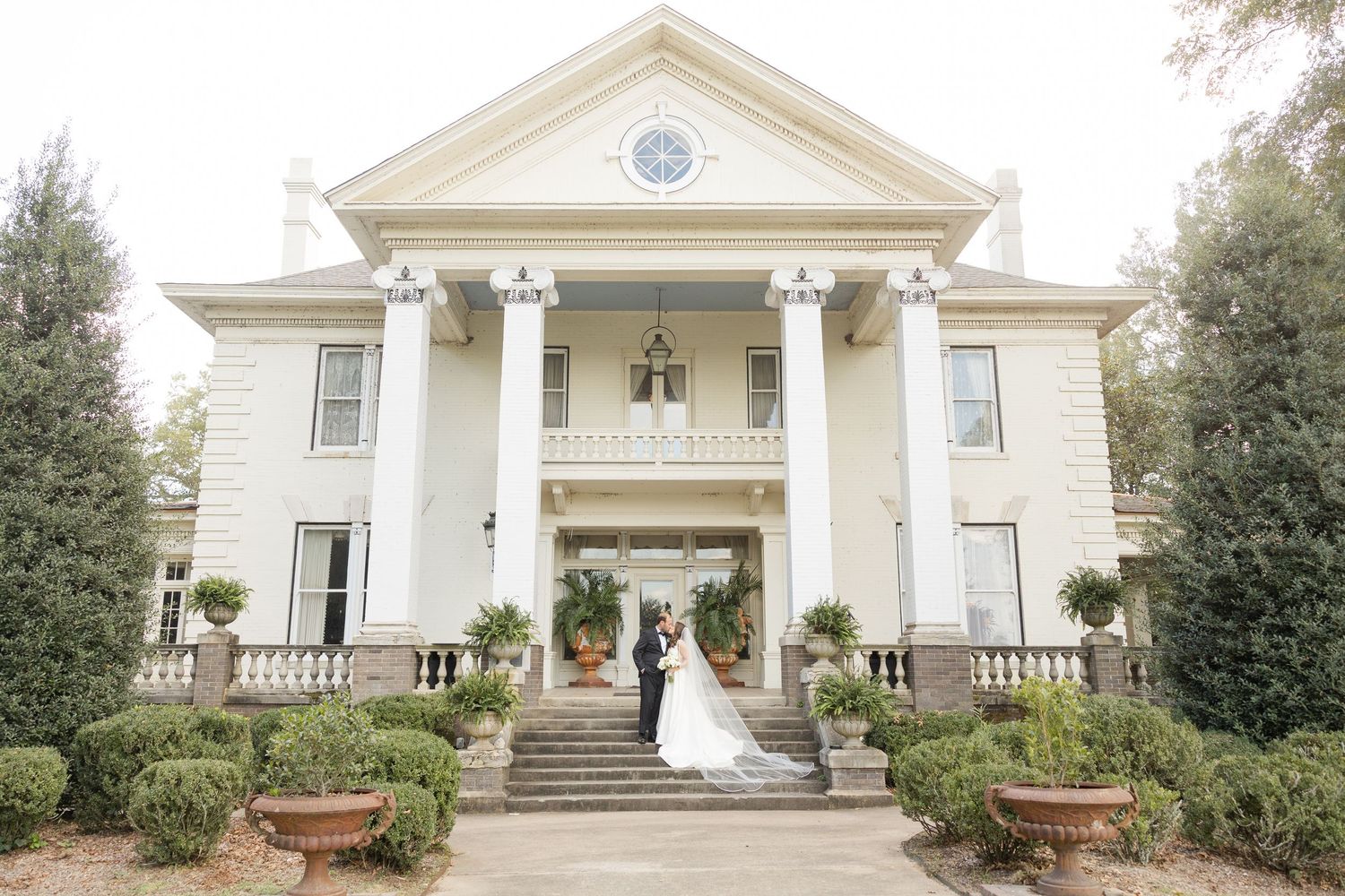 Elegant white mansion with grand columns and stairs features classical Southern architecture and manicured landscaping.
