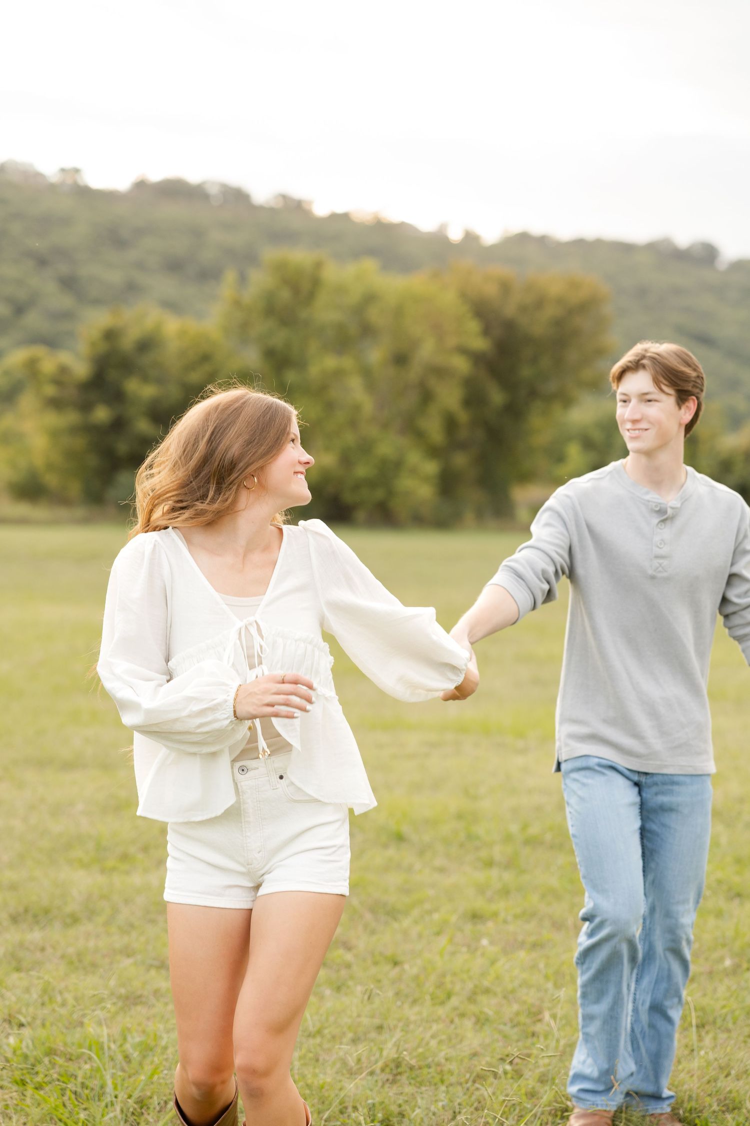 Two people dance and twirl joyfully across a sunlit meadow with trees in the distance.