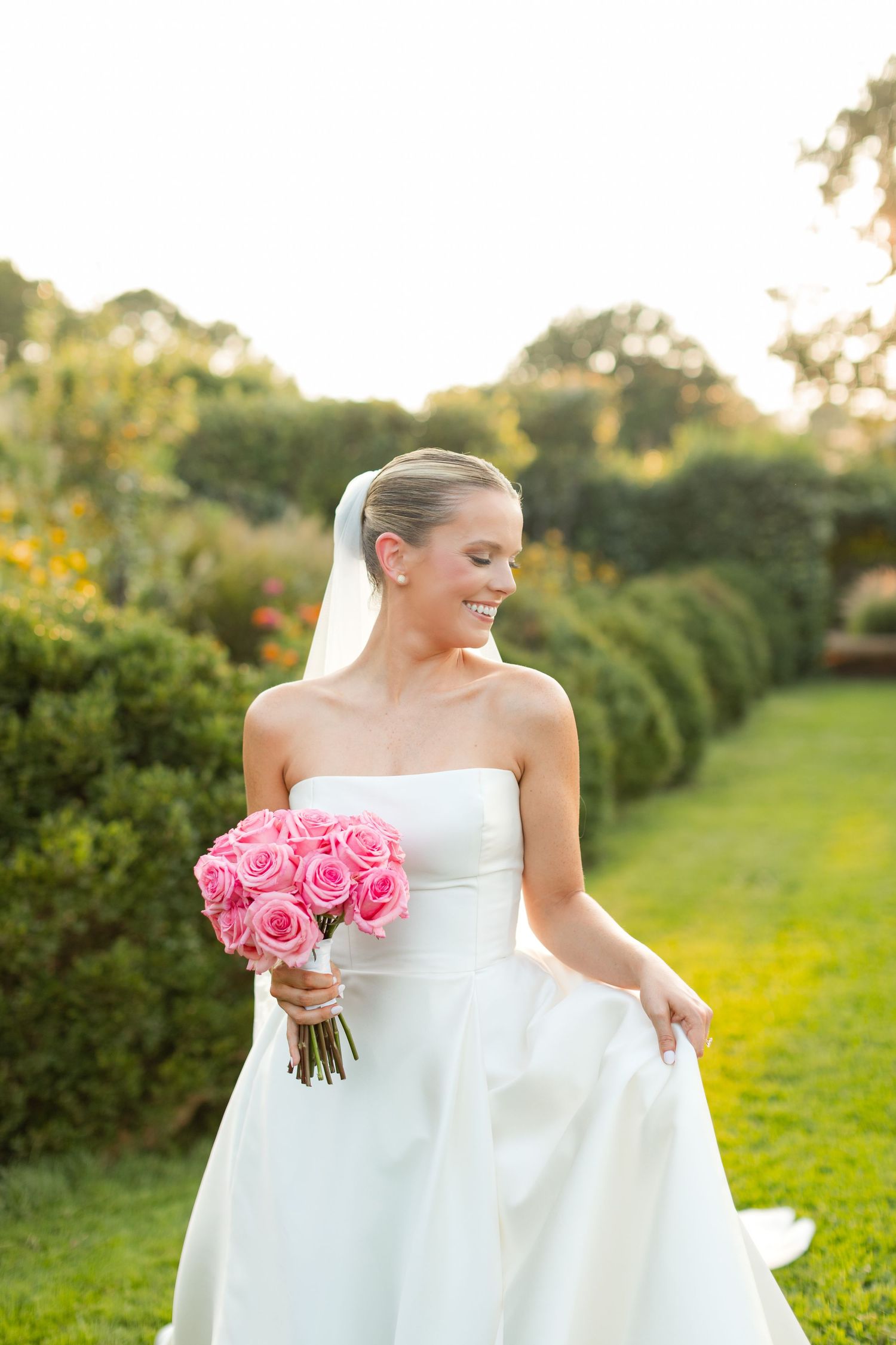 A bride in a white strapless gown holds pink roses while standing in a sunlit garden setting.