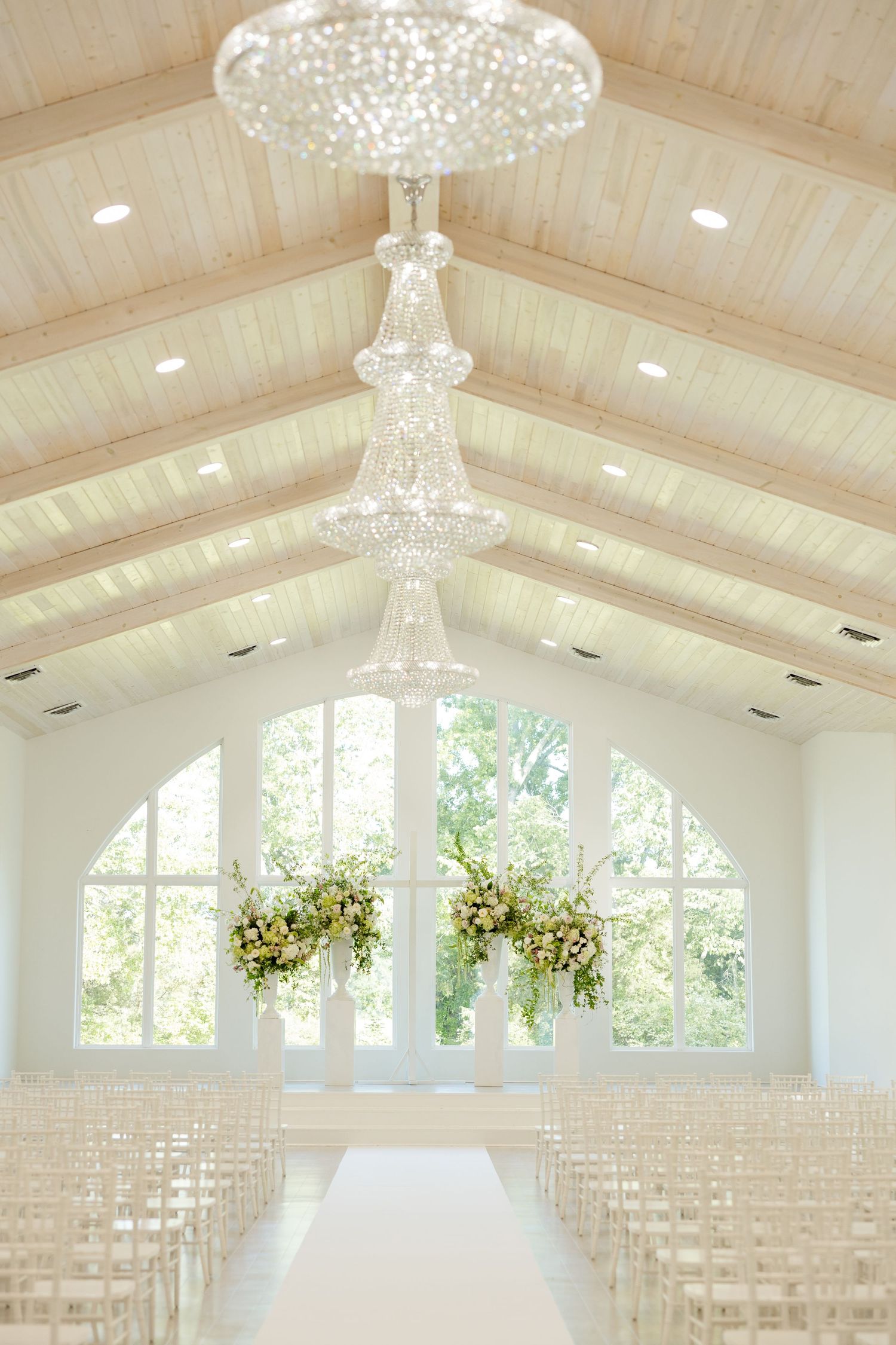 Elegant white wedding chapel with crystal chandeliers, white chairs, and floral arrangements framing a large arched window.