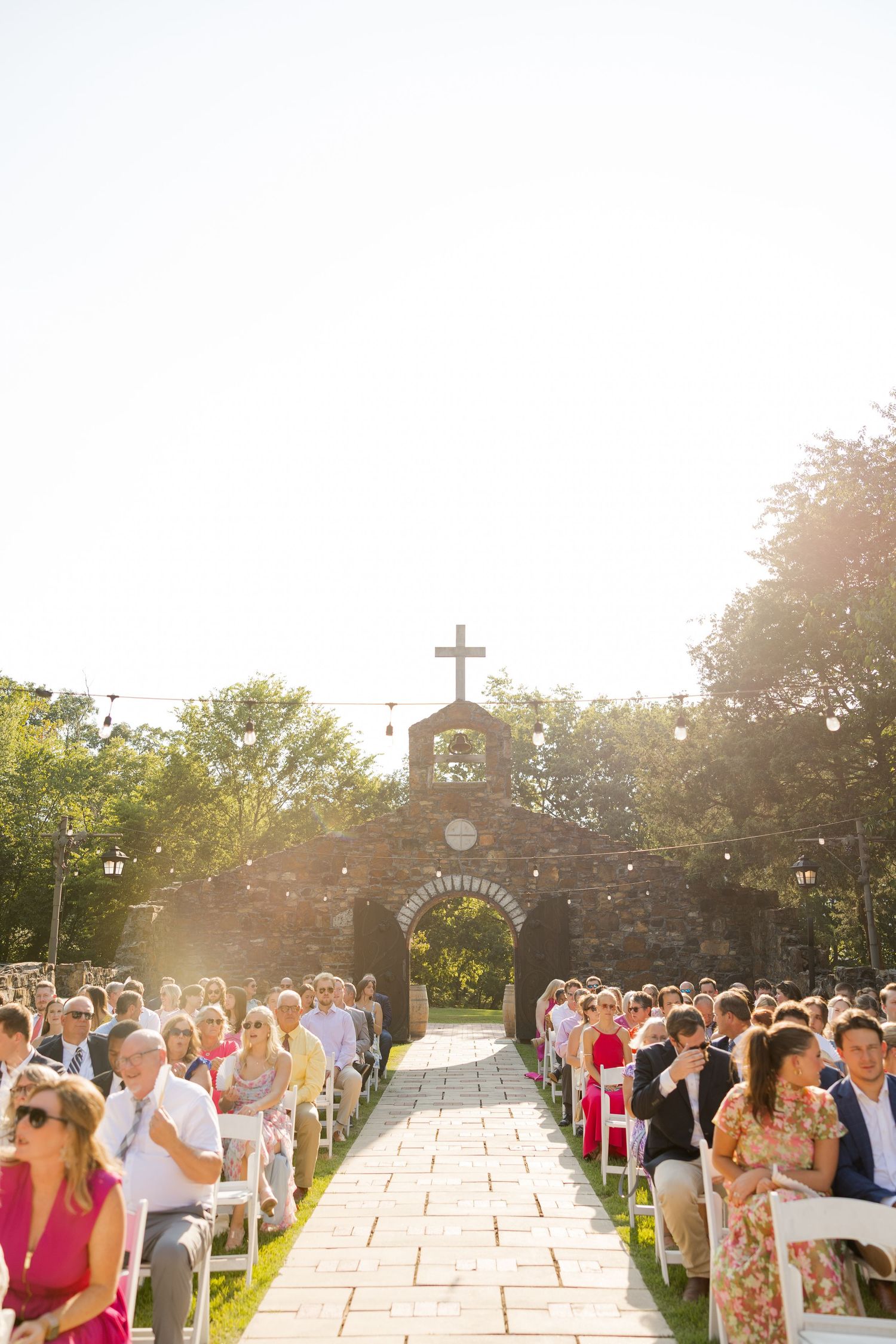 Outdoor wedding ceremony guests stand in rows along a stone pathway during golden hour sunset.