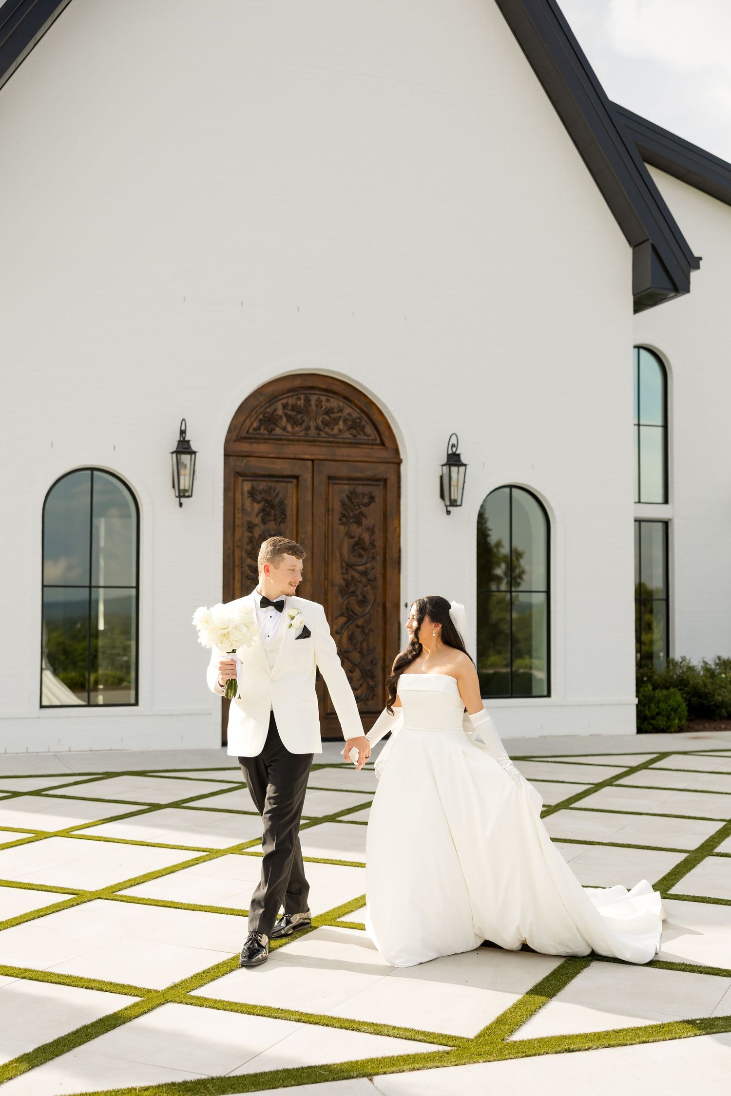 A couple in wedding attire poses in front of a modern white church with geometric grass patterns.