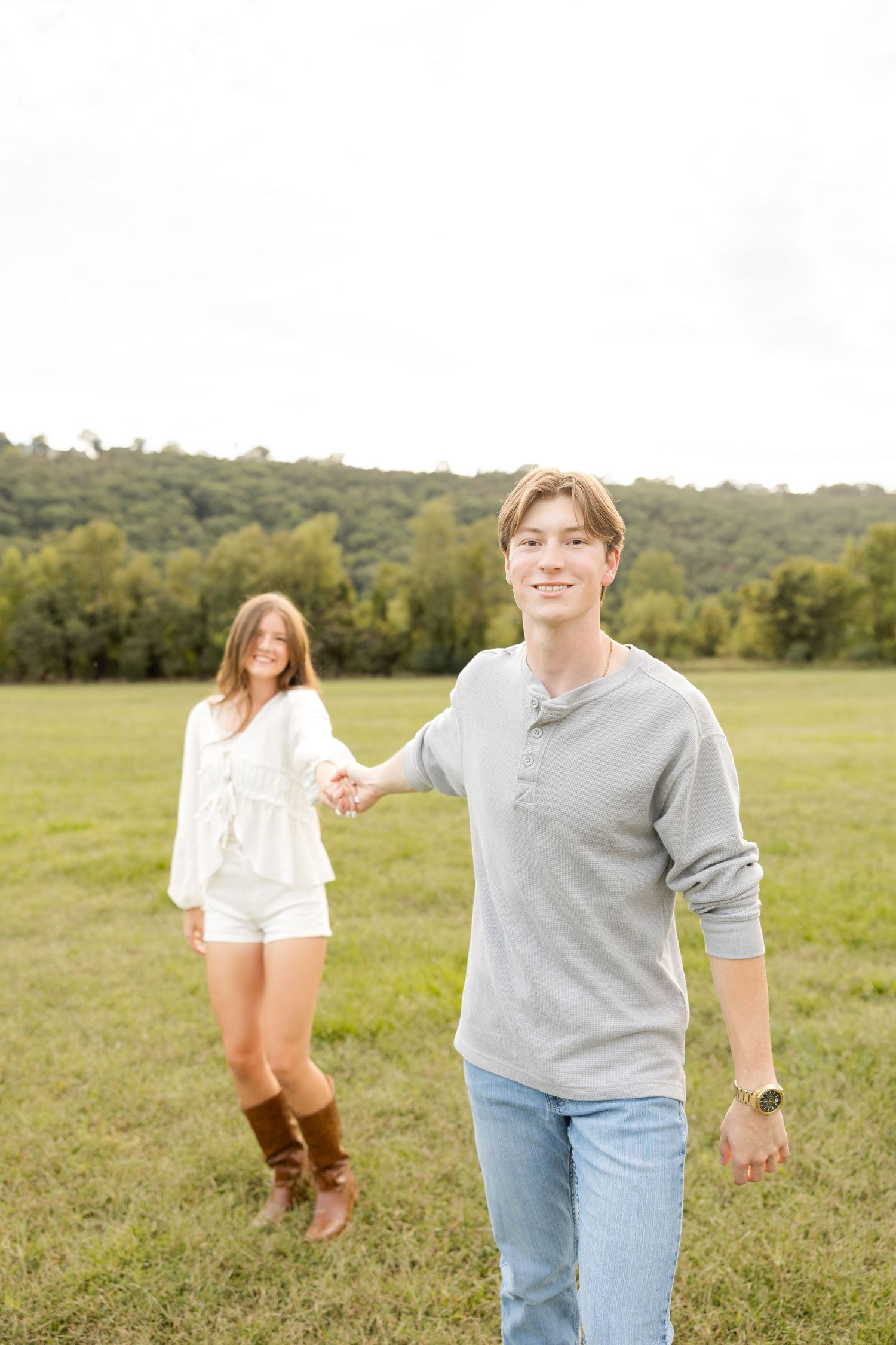 A couple holds hands while walking through a grassy field with hills in the background.