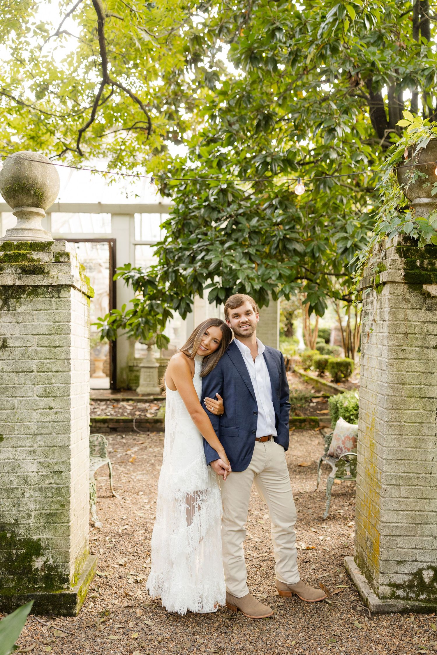 Romantic couple in wedding attire poses between stone pillars in a garden setting.