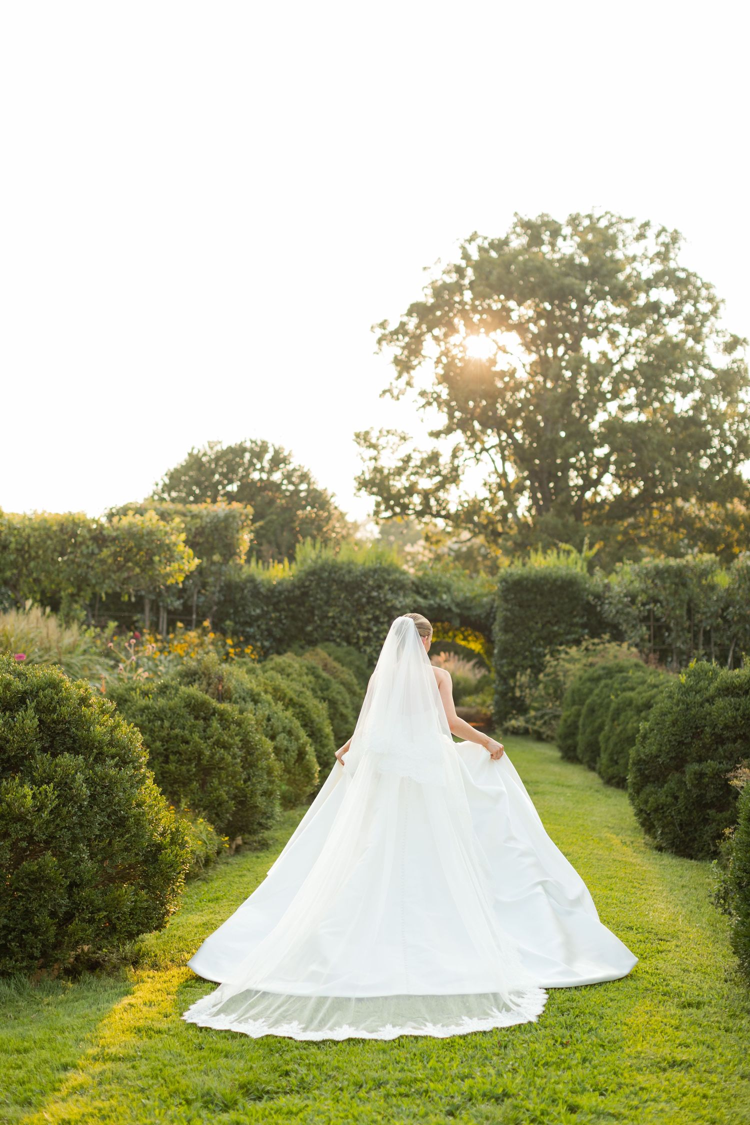 A flowing white wedding dress billows gracefully across a lush green garden path at sunset.