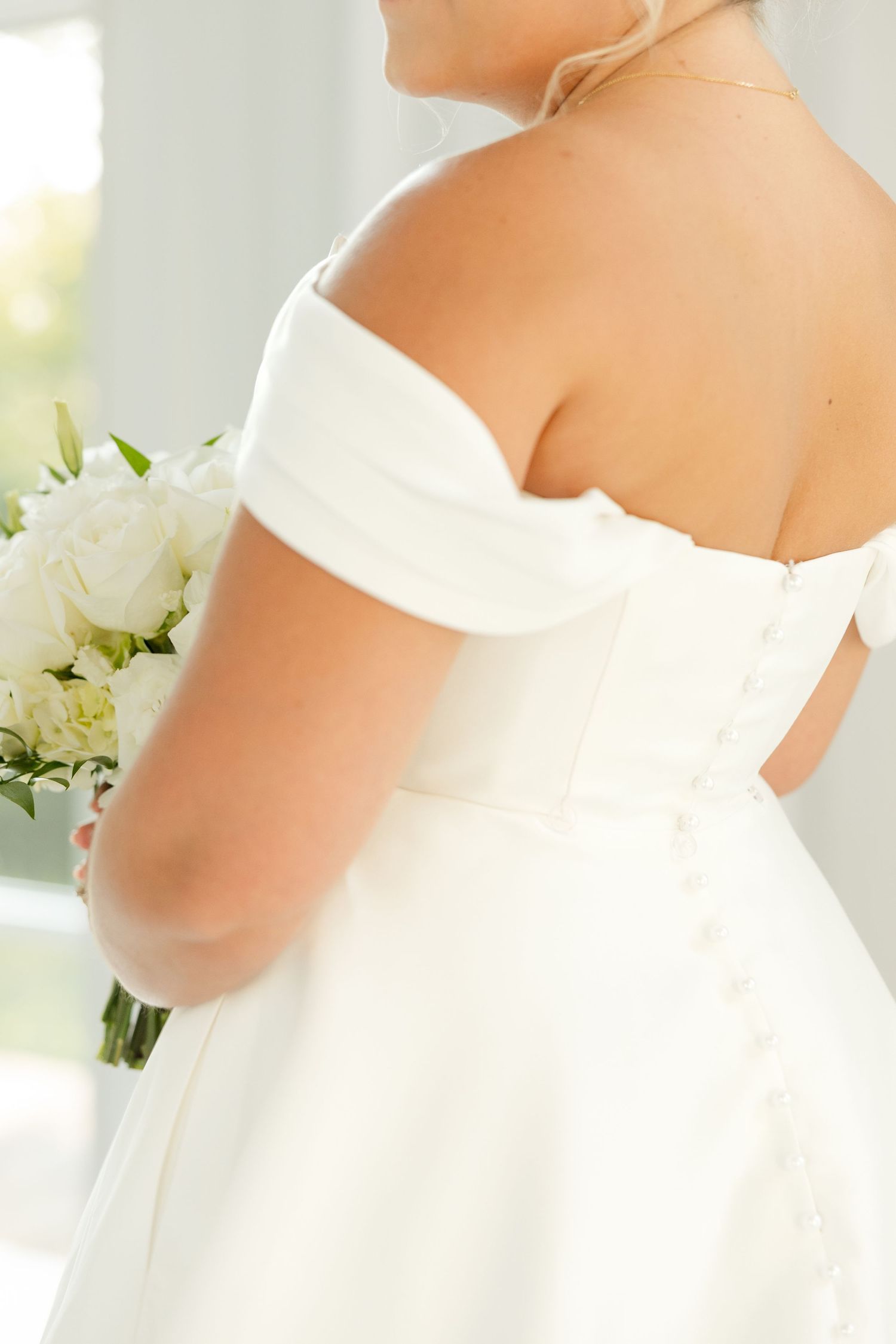Close-up view of a white off-shoulder wedding dress with a bouquet of white flowers against a light background.