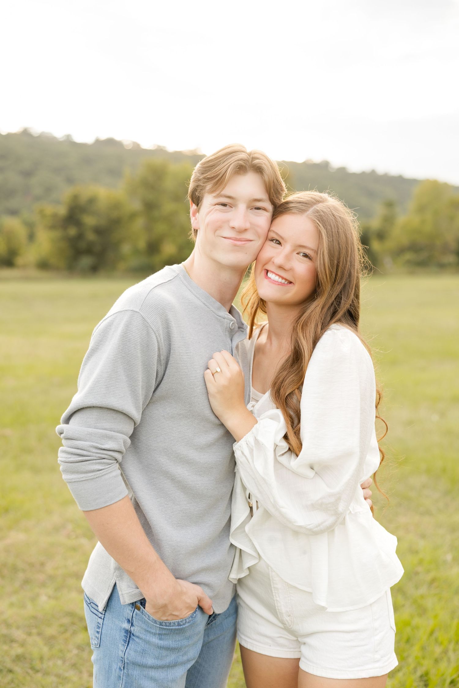 A romantic couple embraces and laughs together in a sunny meadow with rolling hills in the background.