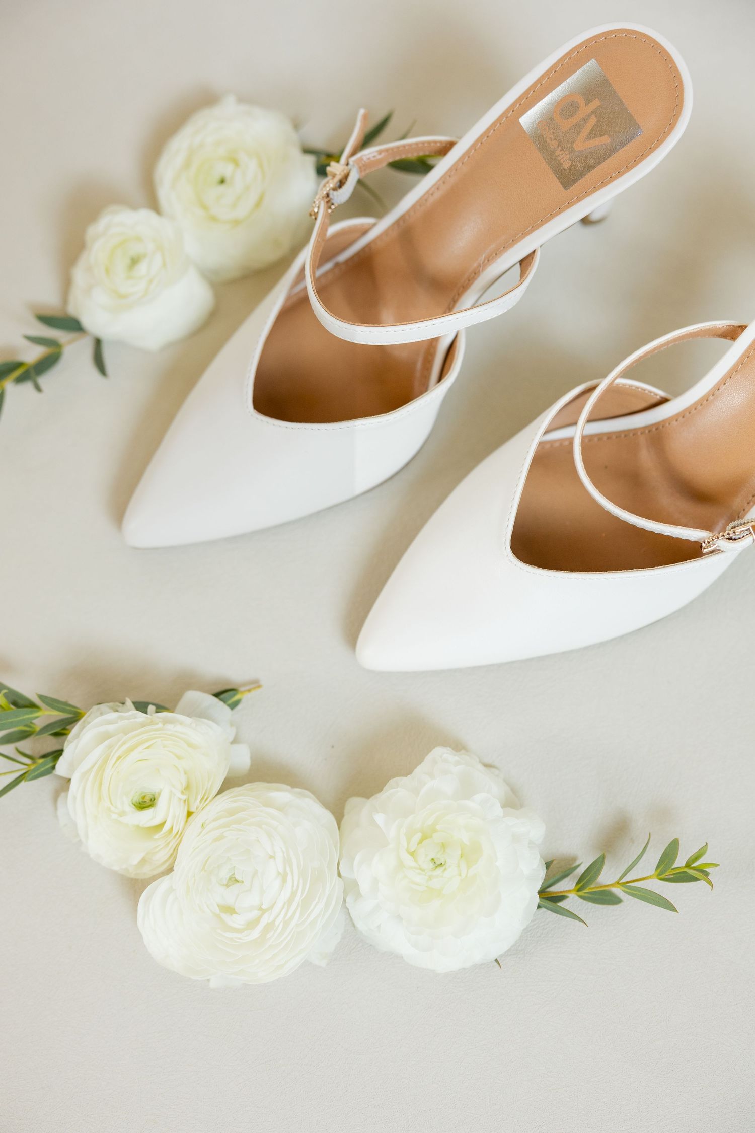White bridal shoes and fresh white ranunculus flowers arranged on a light beige surface.