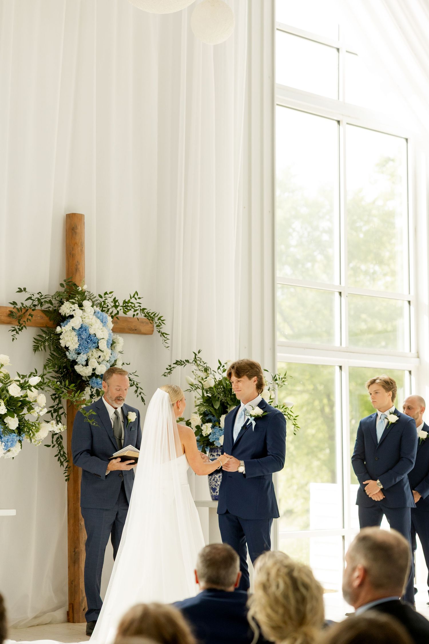 Wedding ceremony in progress inside a bright white chapel with wooden cross and floral arrangements.Cold Springs Events
