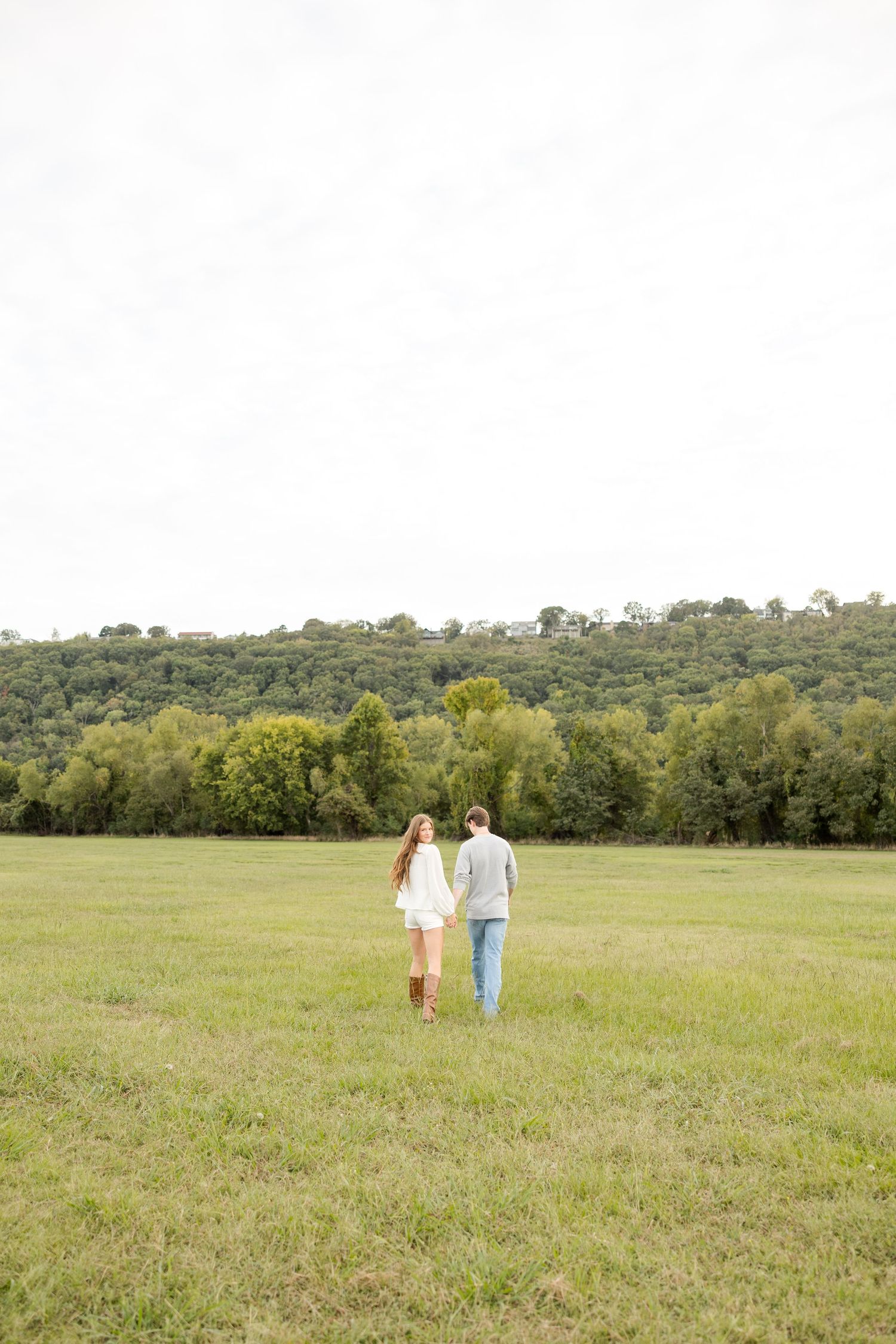 A couple walks hand in hand through a green meadow with rolling hills in the background.