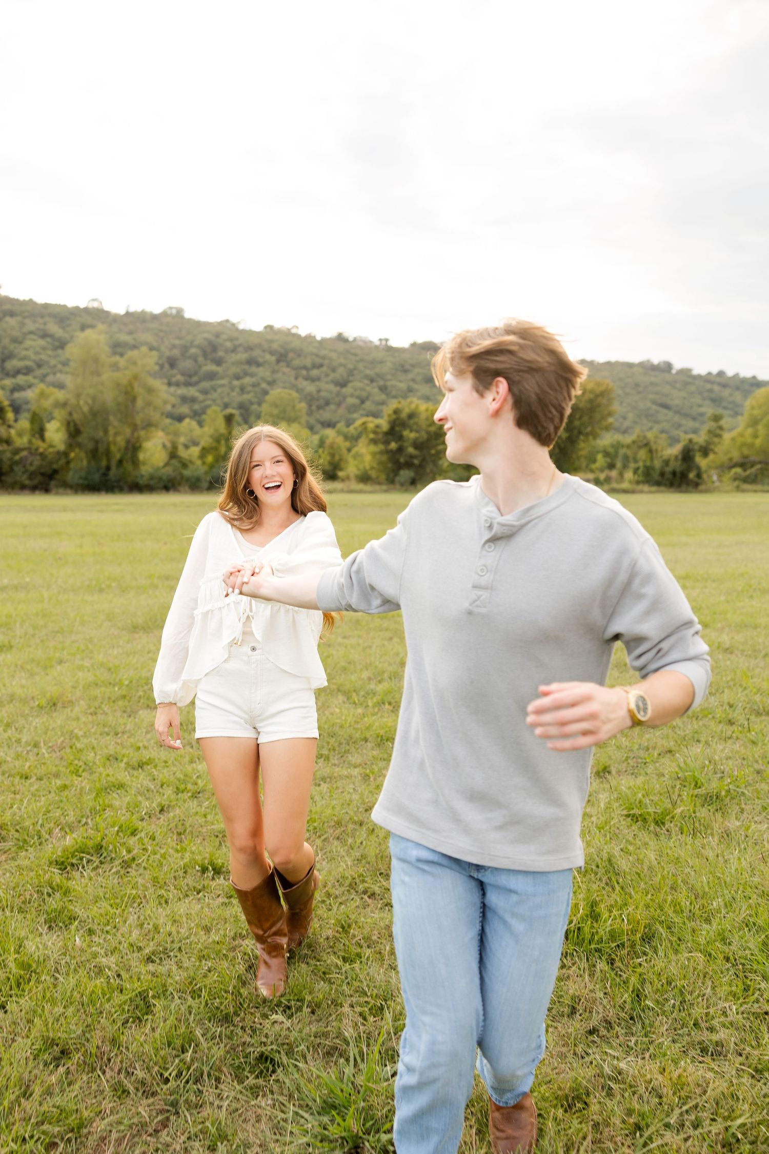 Two people laugh and dance together in a sunny green field with hills in the background.