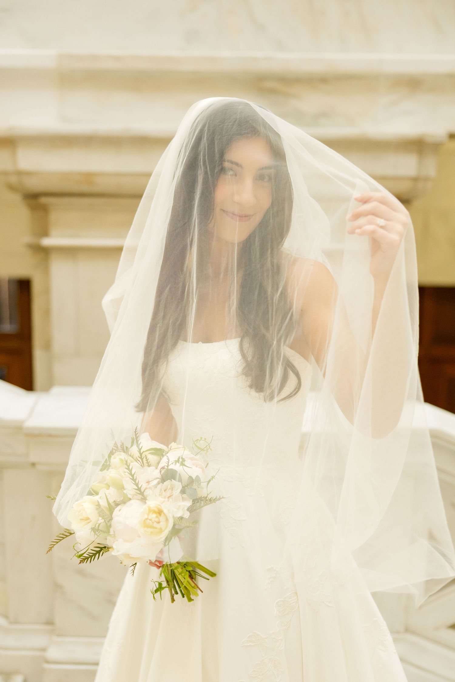 A bride in a white wedding dress holds a bouquet of cream roses while wearing a flowing veil in an elegant setting.