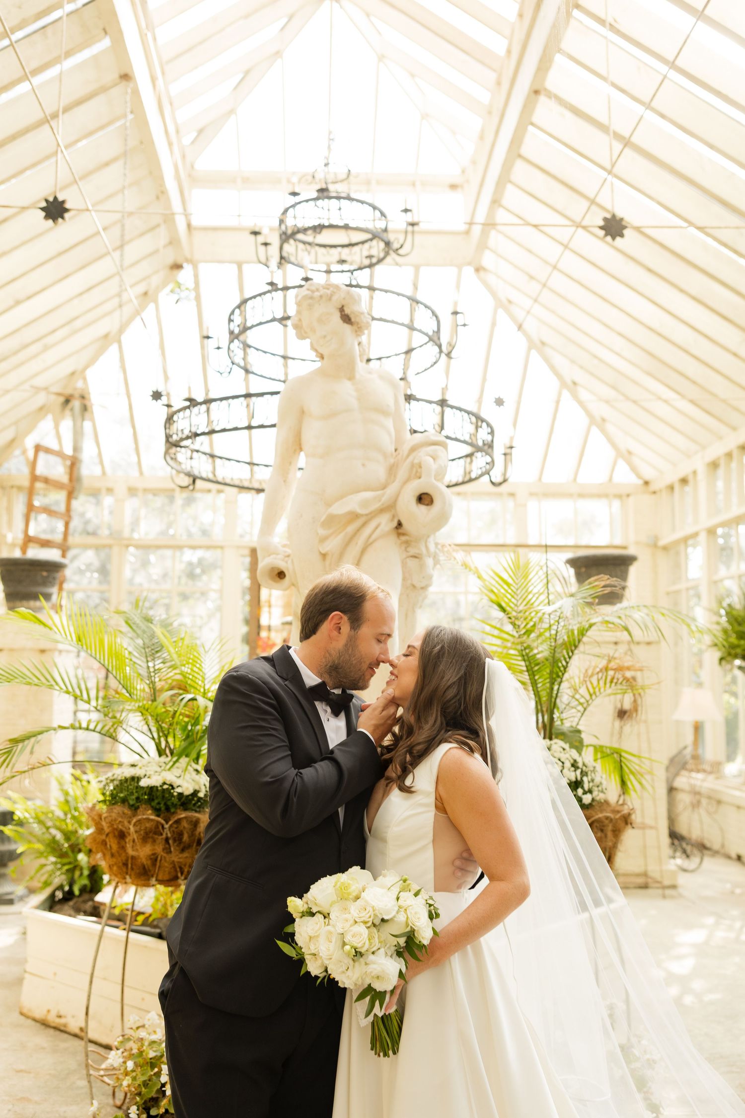A romantic wedding couple shares a kiss in an elegant greenhouse conservatory filled with tropical plants and chandeliers.