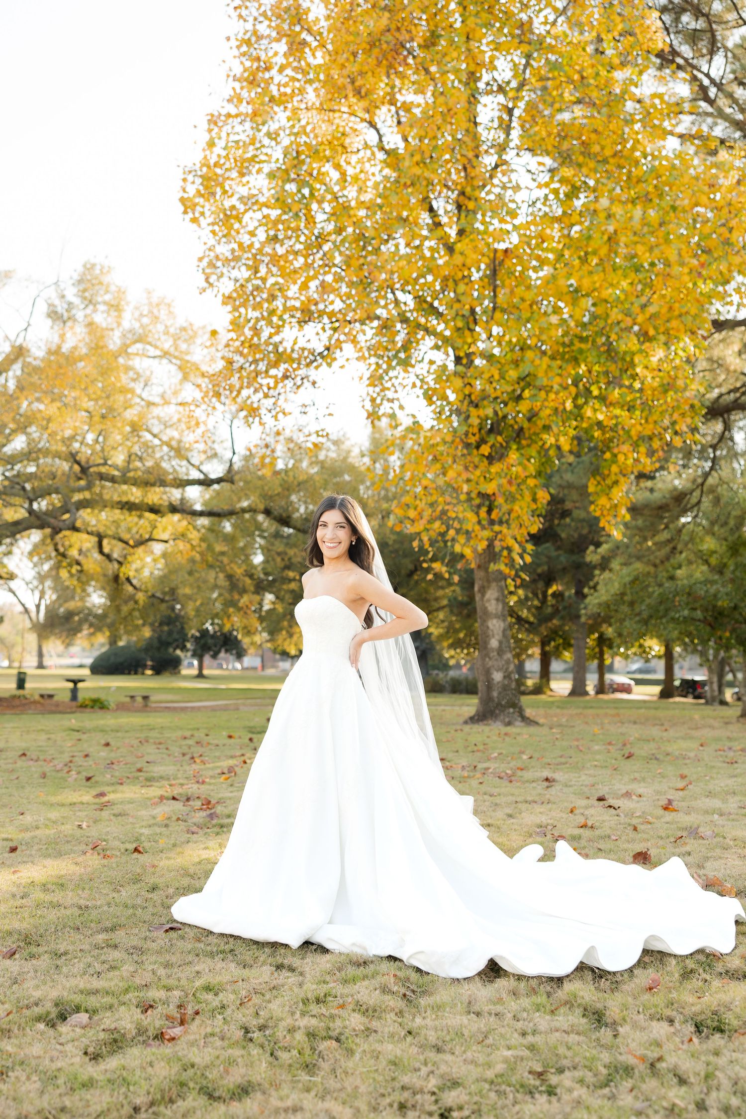 A flowing white wedding dress spreads across autumn grass with golden trees in the background.