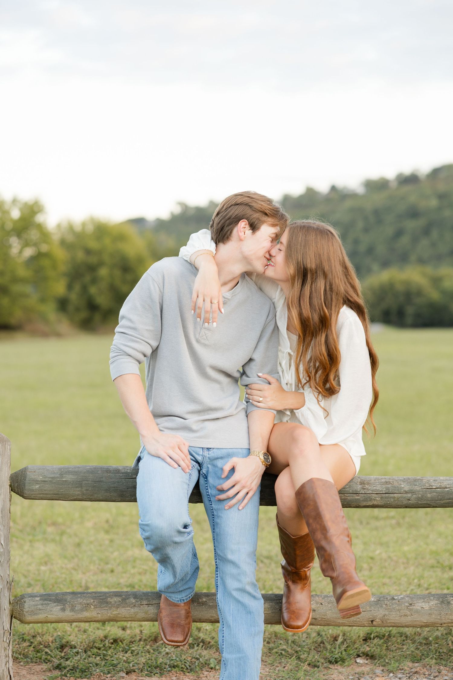 A couple sitting together on a wooden fence in a pastoral field setting.
