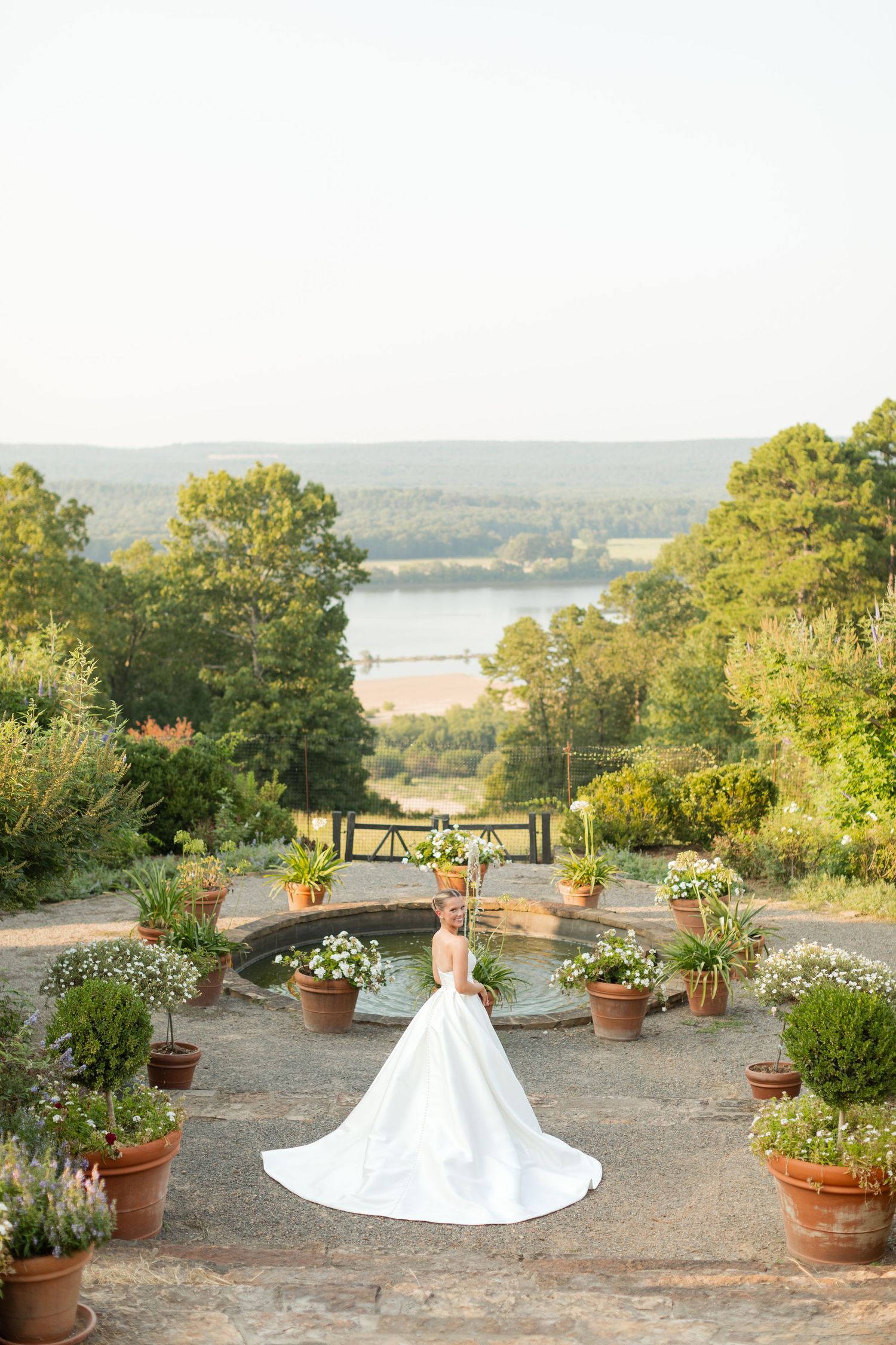 A bride in a flowing white gown stands on a garden terrace overlooking a scenic lake view surrounded by potted plants.