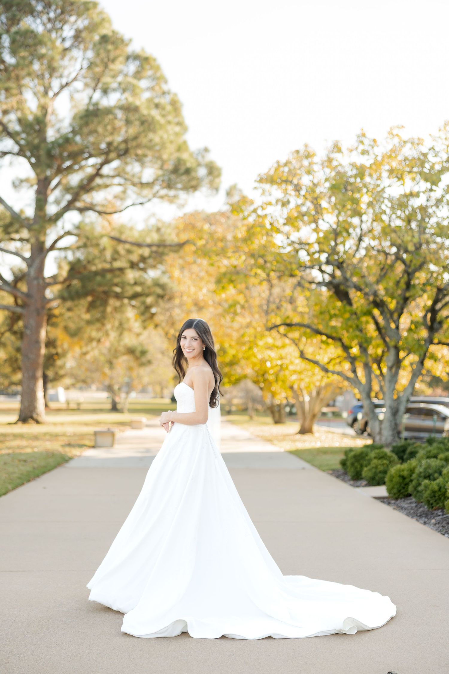 An elegant white wedding dress flows across a path surrounded by autumn trees and golden foliage.