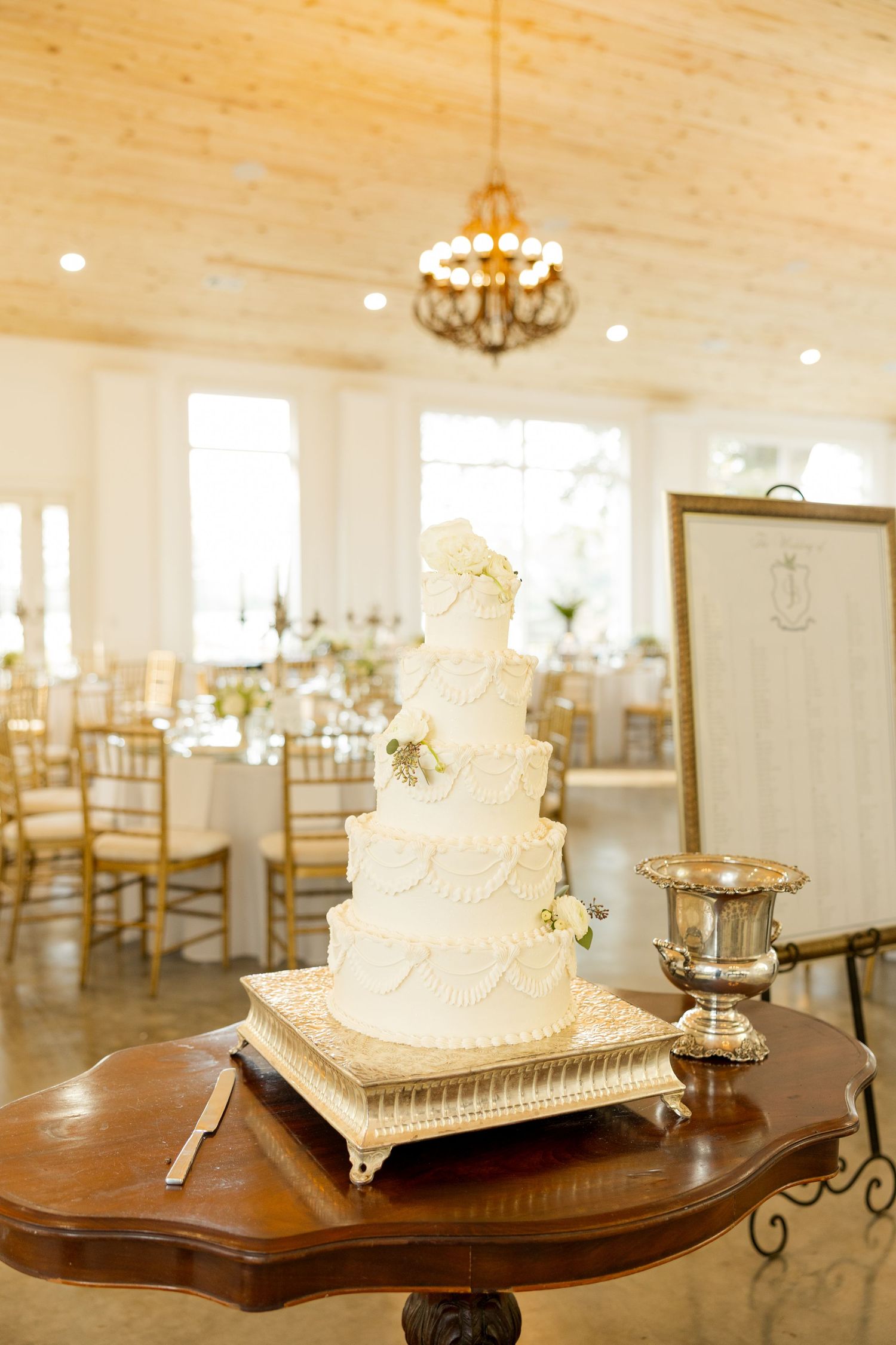 Elegant white wedding cake displayed on wooden table in bright reception hall with chandeliers.Marlgsate Plantation