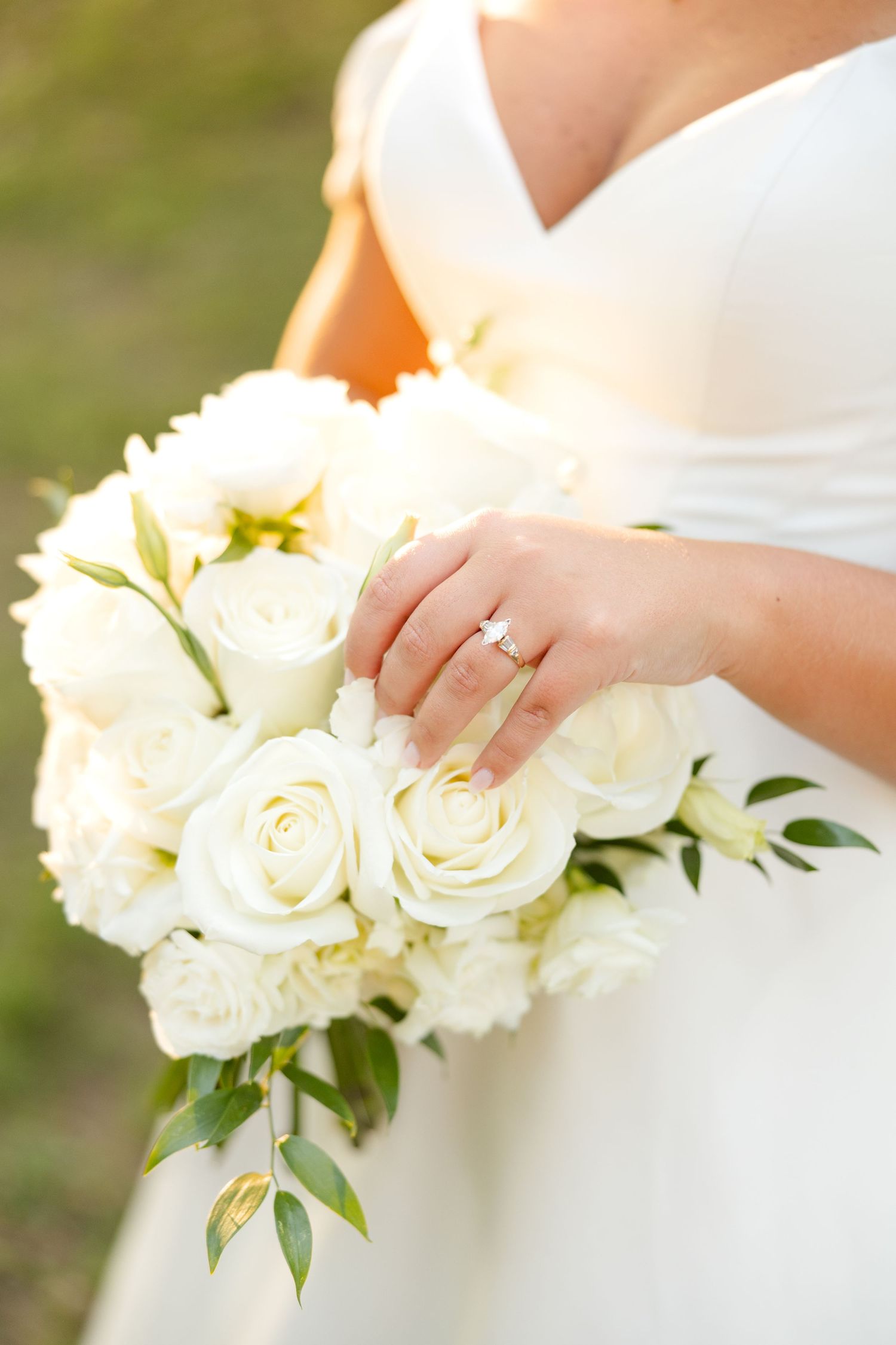 Close-up shots of white rose bridal bouquet with diamond engagement ring on bride's hand in natural lighting.