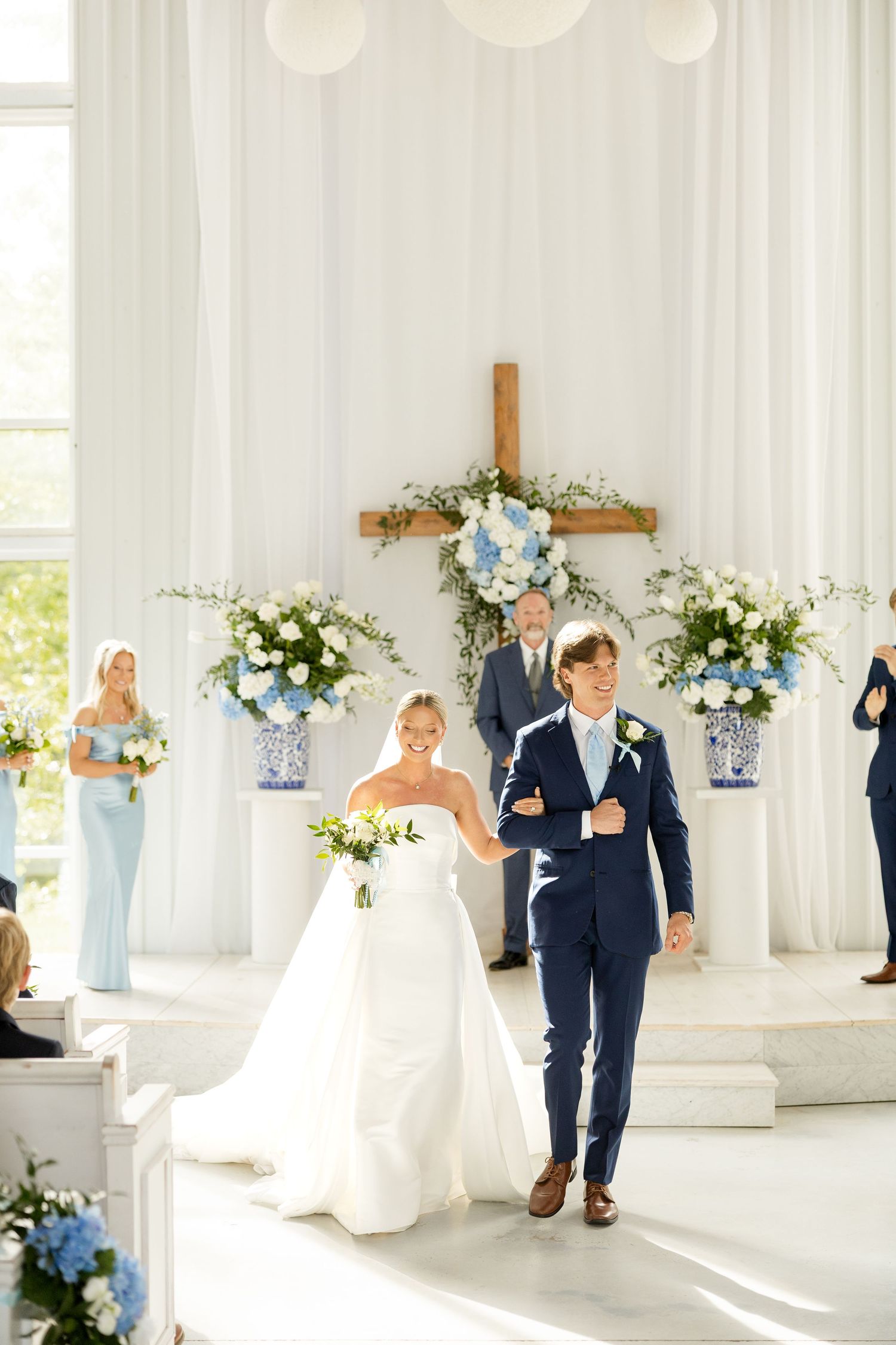 Elegant wedding ceremony in a bright white indoor venue decorated with blue and white floral arrangements and crosses. Cold Springs