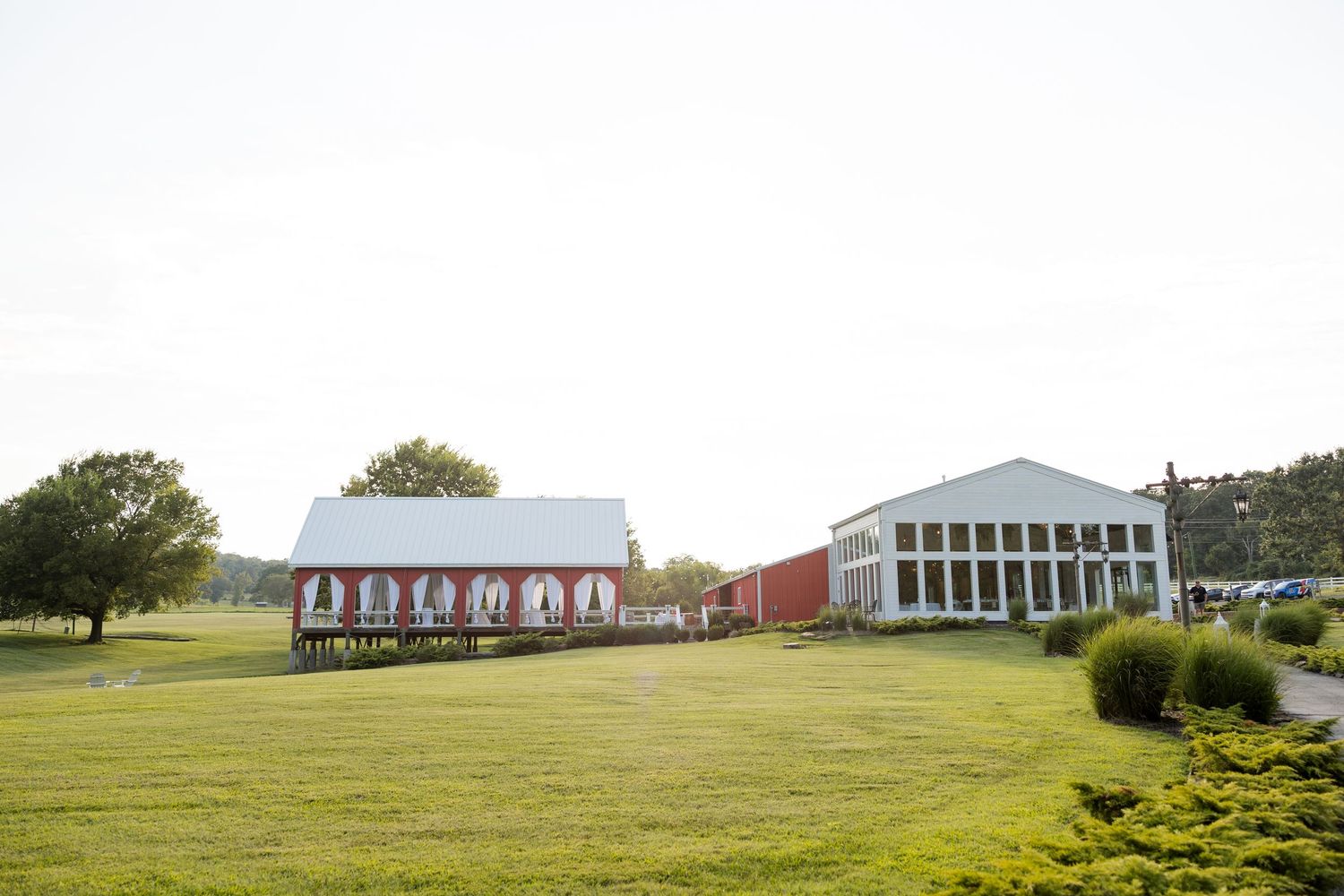 A red barn and white event venue building sit on a grassy hillside surrounded by trees.