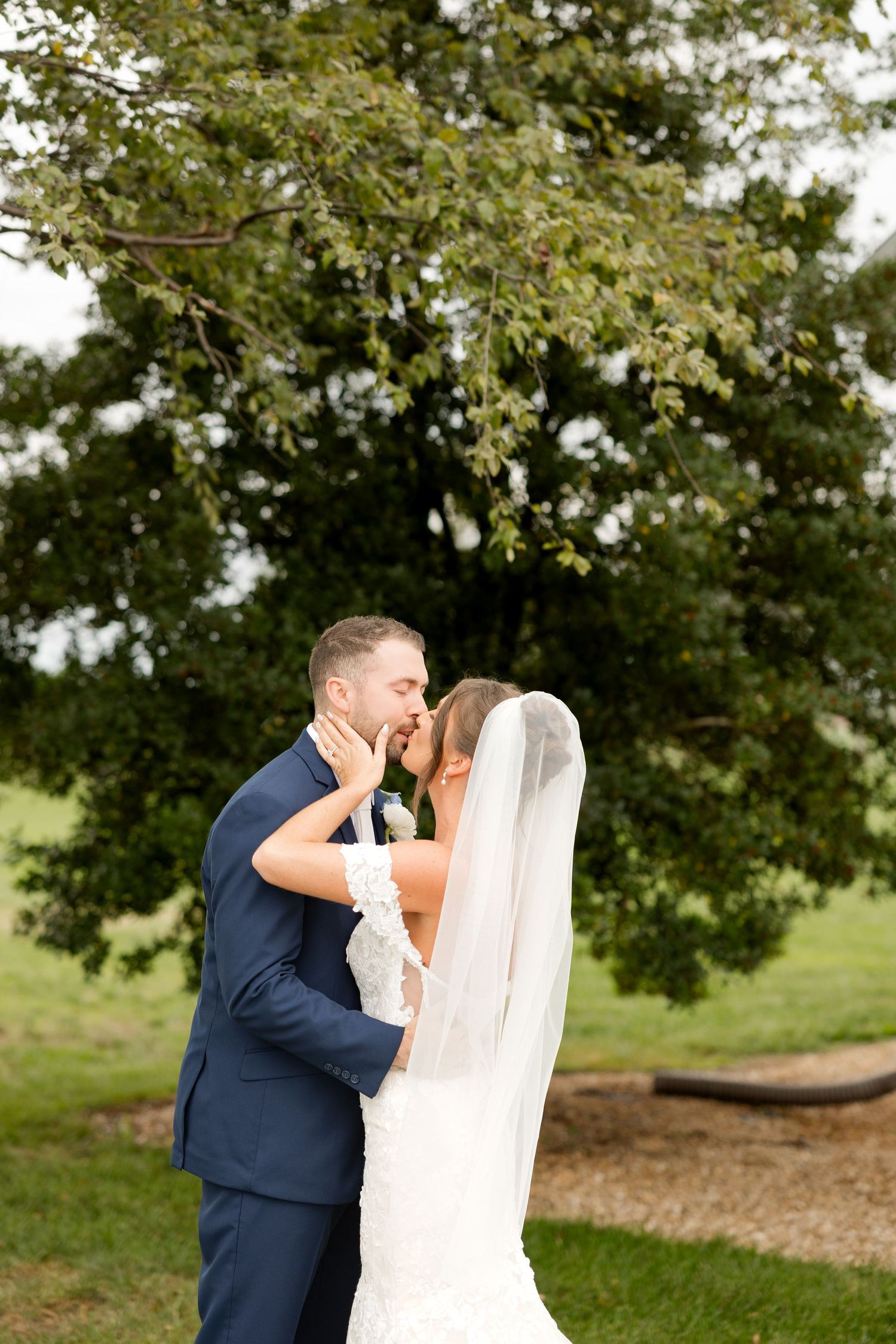 A romantic wedding couple embraces outdoors under a large tree during their special day.
