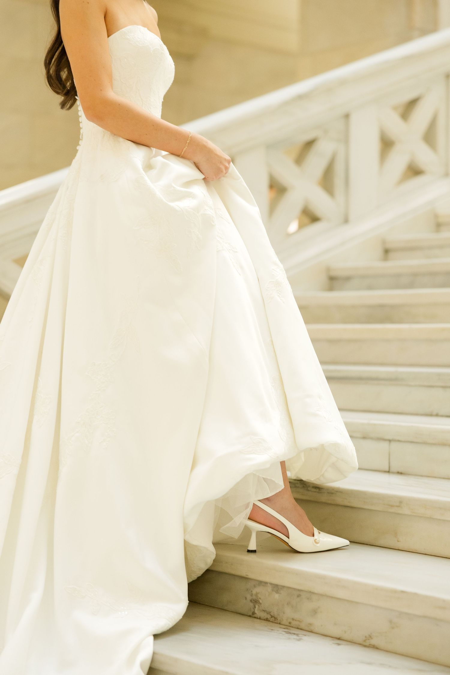 A bride in a flowing white wedding dress walks up elegant marble stairs while holding the skirt of her gown.