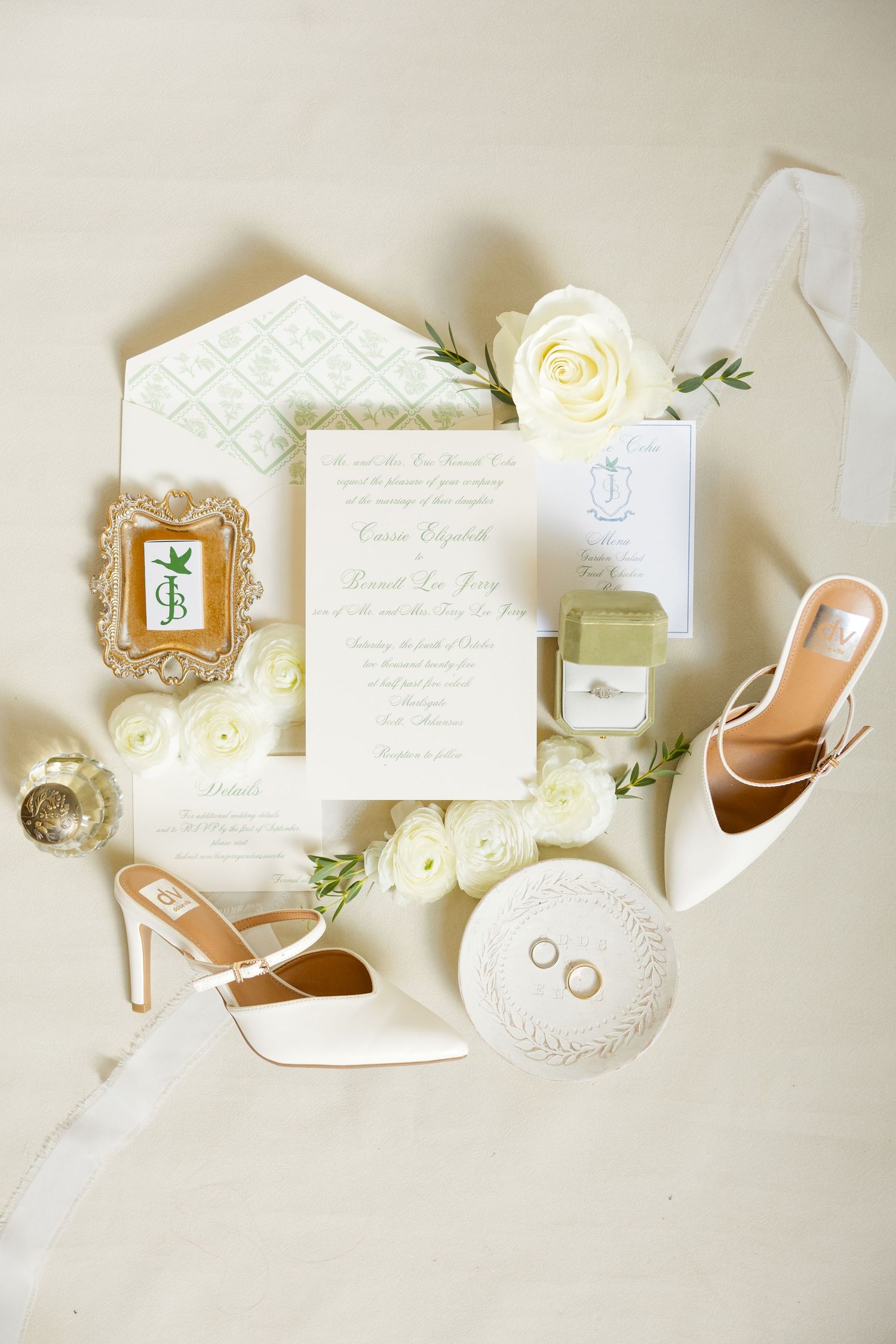Elegant wedding details flatlay with white invitation, roses, heels, rings and decorative frames on cream background.