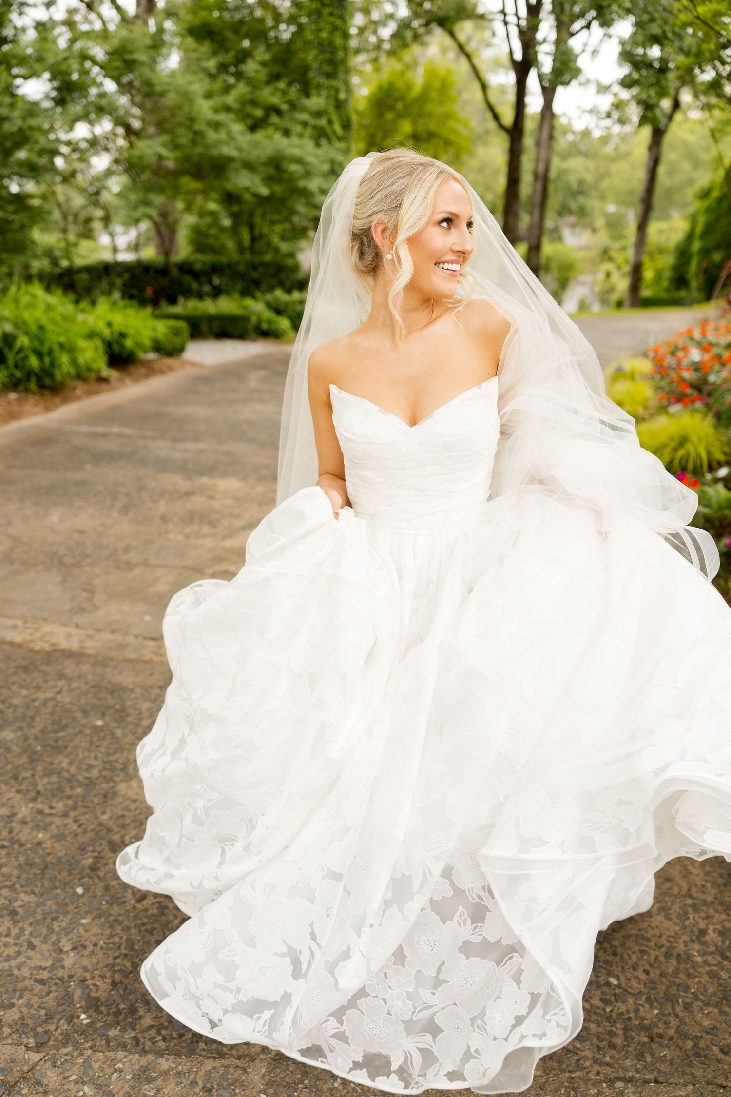 A bride in a flowing white wedding dress twirls gracefully on a garden path surrounded by lush greenery.