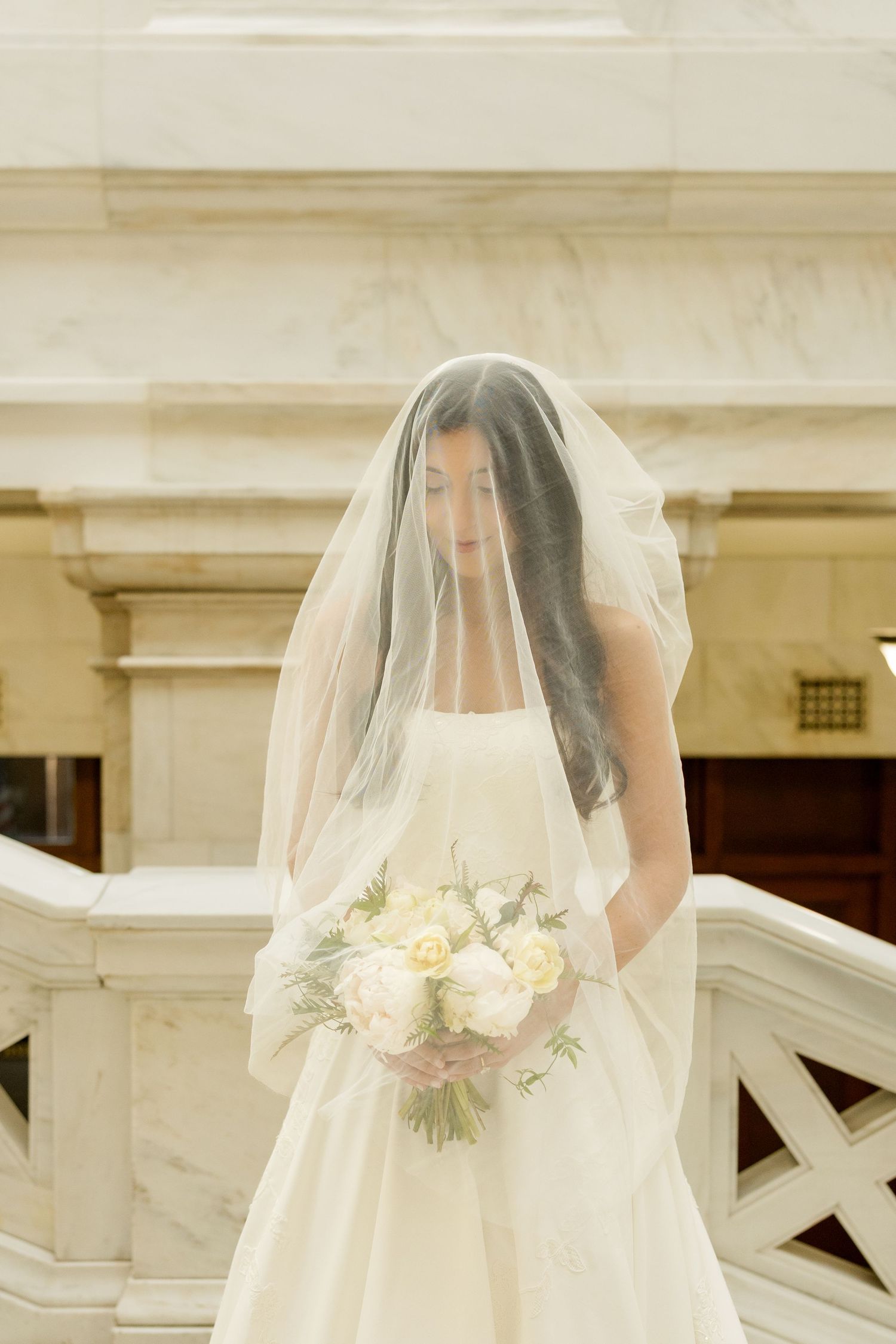 A bride in a flowing white gown holds a delicate bouquet of white flowers against a marble architectural backdrop.