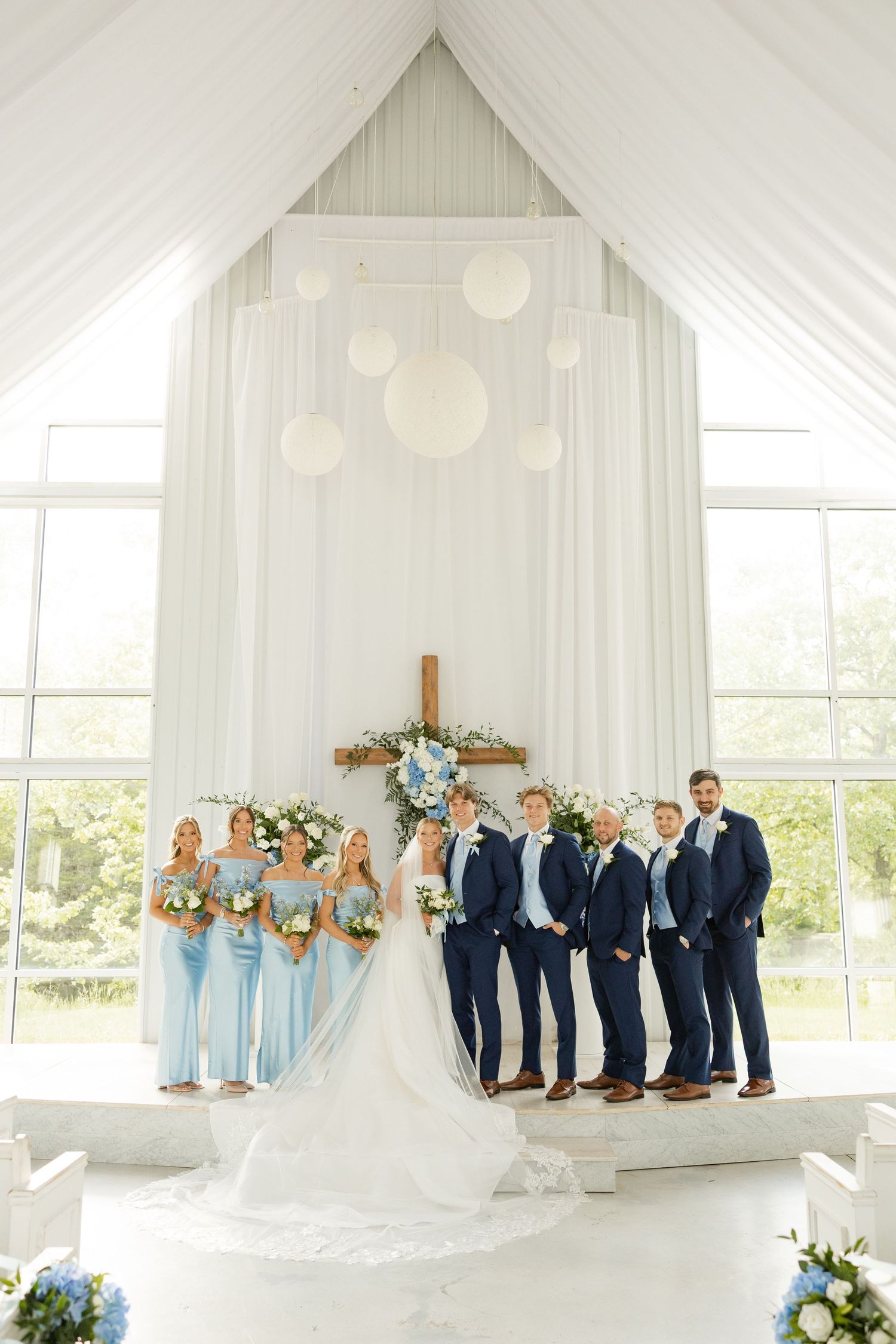 Wedding party poses in a modern white chapel with hanging lanterns and floral cross backdrop.Cold Springs Events