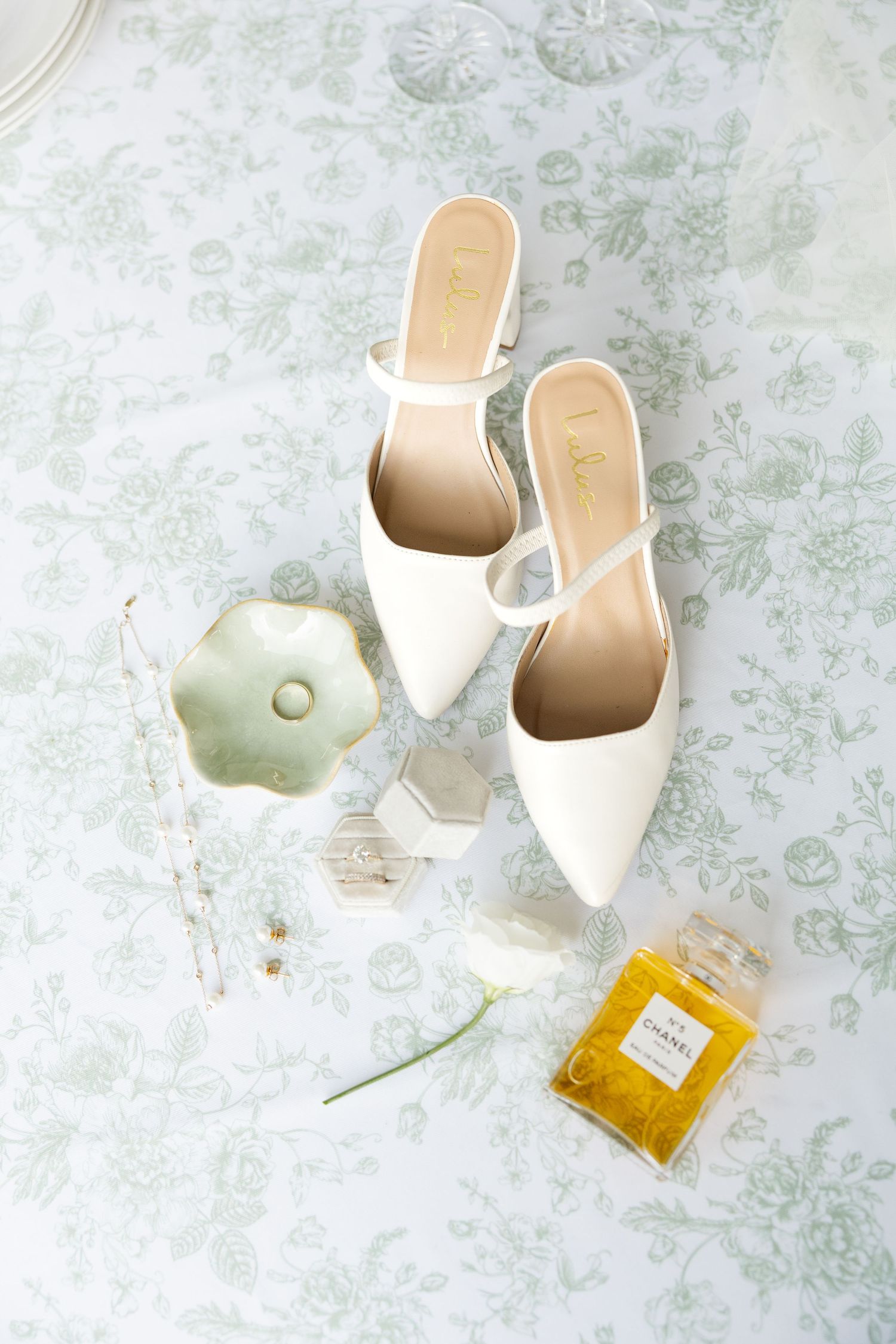Wedding accessories including white heels and perfume bottles arranged on a floral patterned surface.Botanical Gardens of the Ozarks