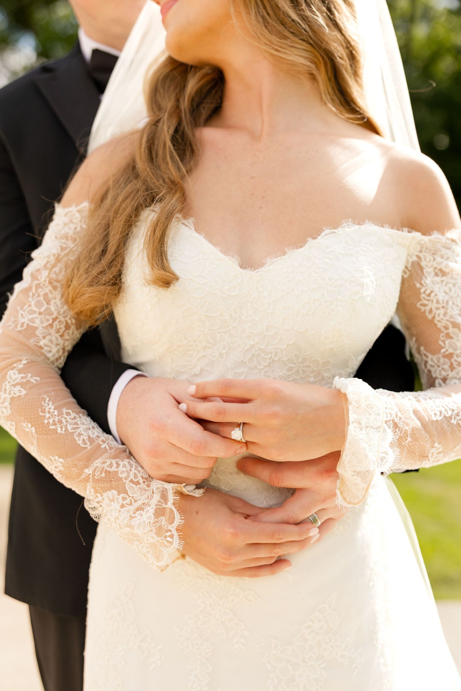 Bride in off-shoulder lace wedding gown holds hands with groom in black tuxedo outdoors.