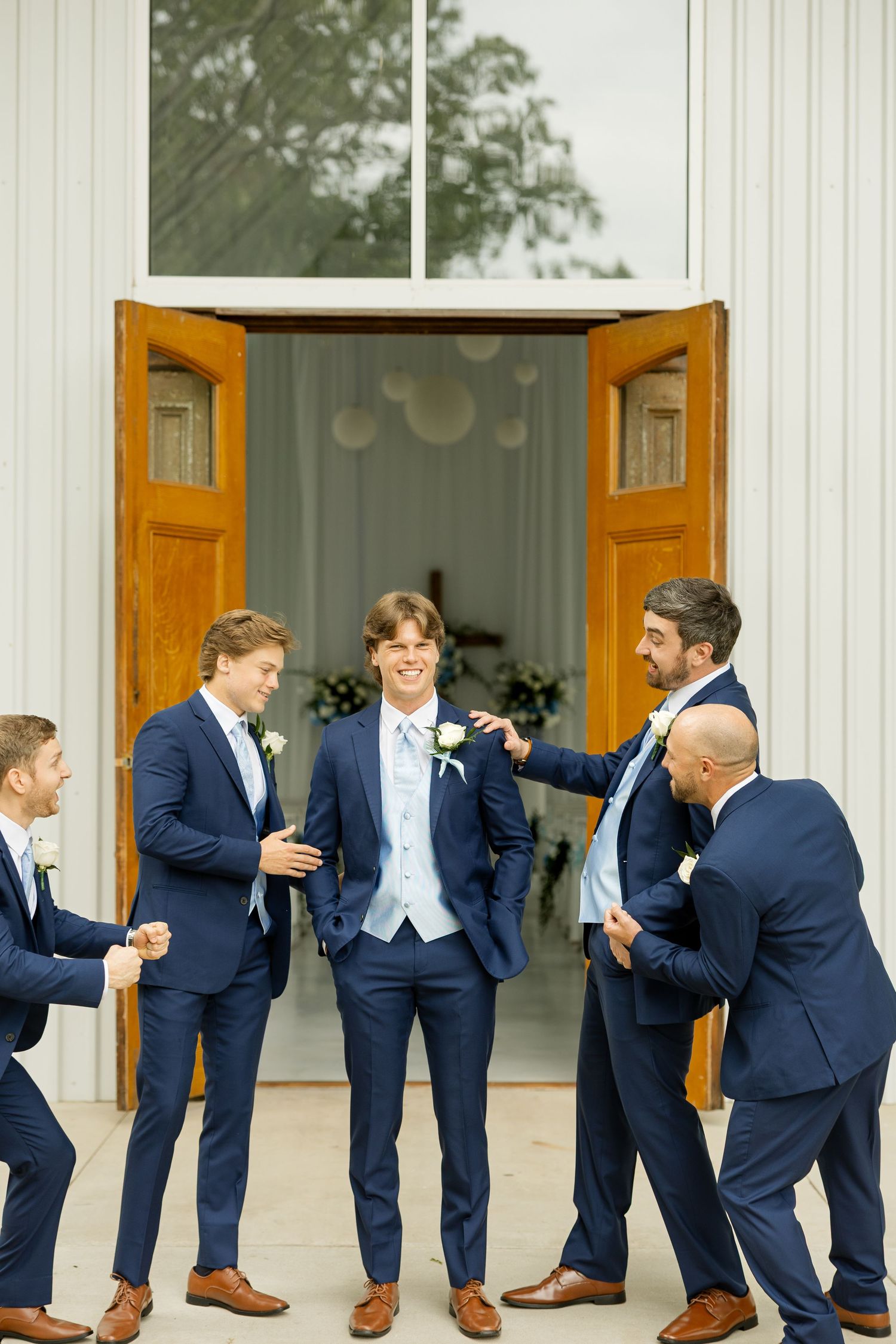 Groomsmen in navy suits striking playful poses outside a white chapel.Cold Springs Events