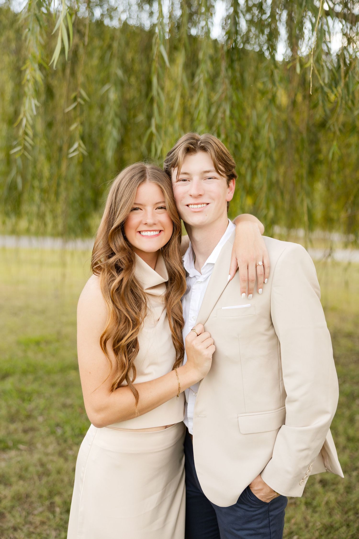 Two people in coordinating cream outfits laugh together beneath drooping willow branches on a sunny day.