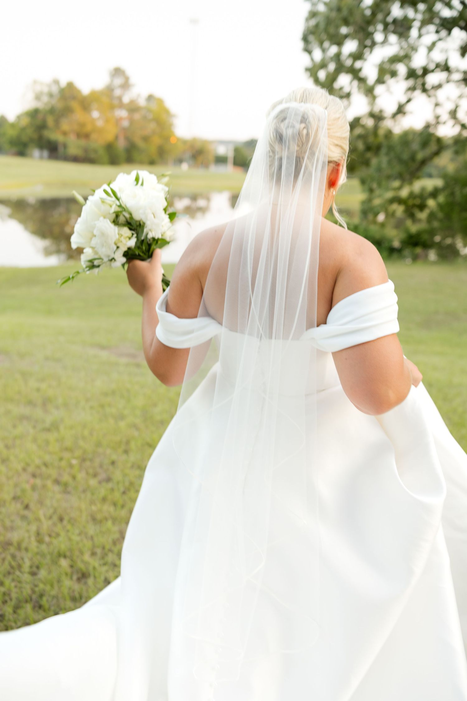 Back view of an off-shoulder wedding gown with delicate veil and white bouquet.