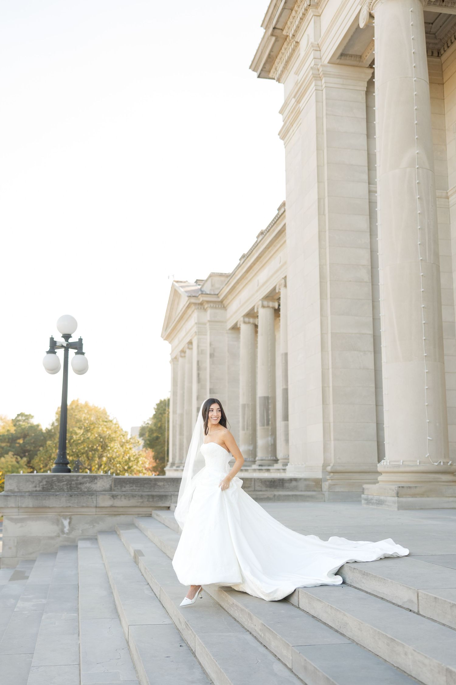 A bride in a white gown with flowing train poses among grand marble columns outdoors.